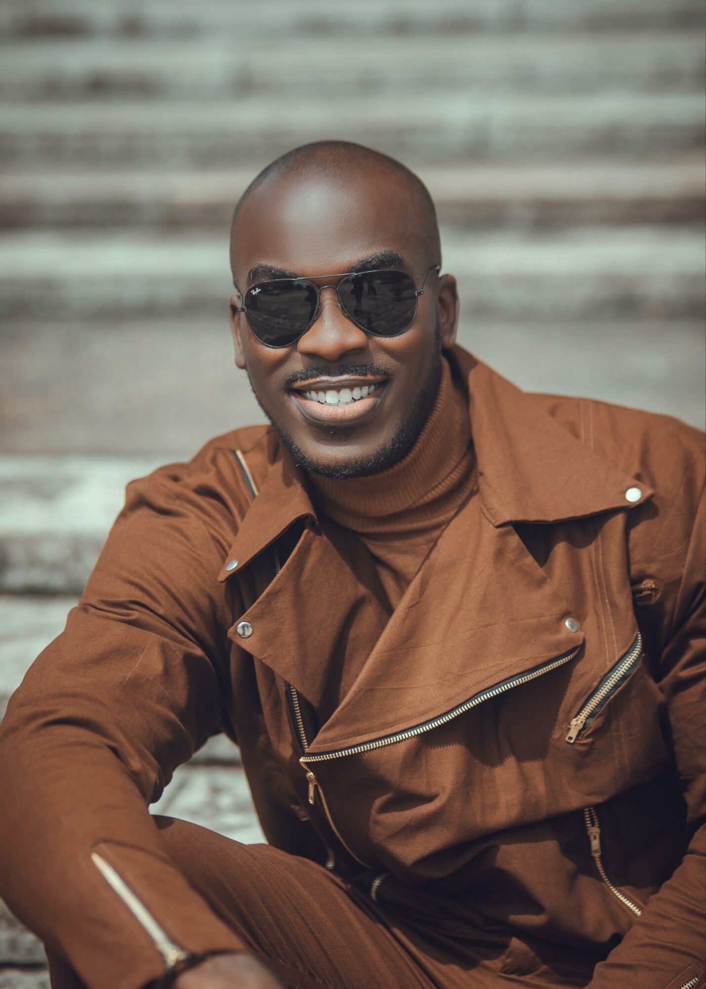 Man wearing sunglasses and a brown jacket, smiling outdoors with a blurred background.