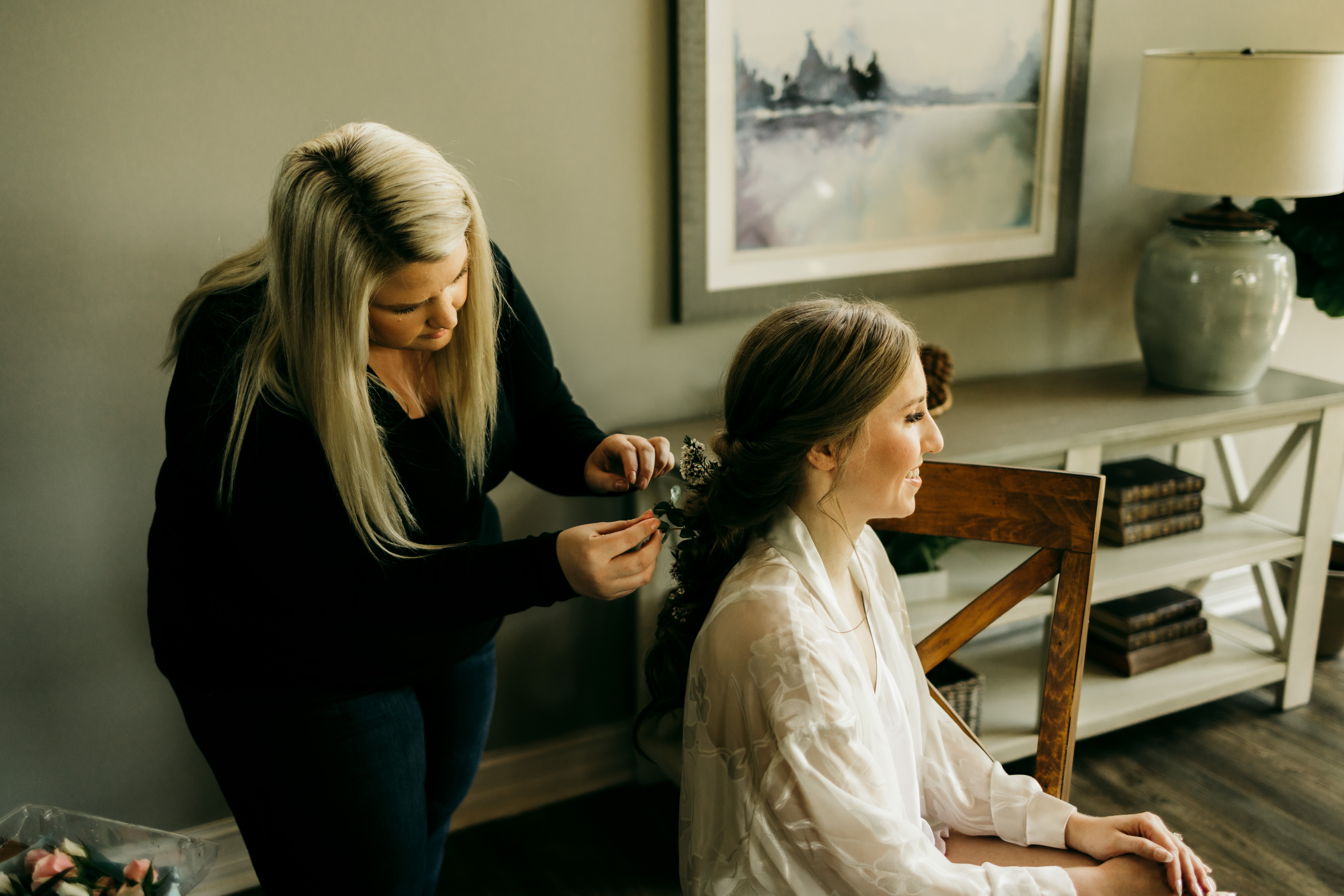 A woman with long blonde hair styles a seated woman with dark hair in a white robe, in a room with artwork and a shelf.