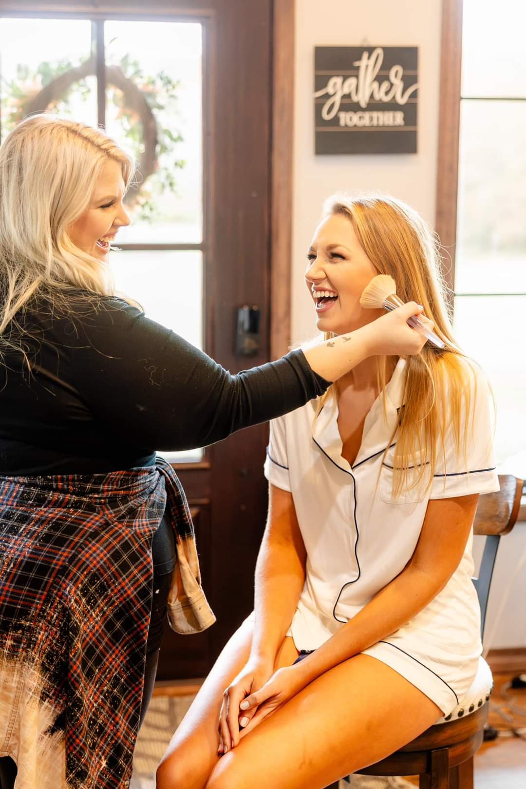 Two women smiling and laughing indoors, one applying makeup to the other, with a window and wall decor in the background.