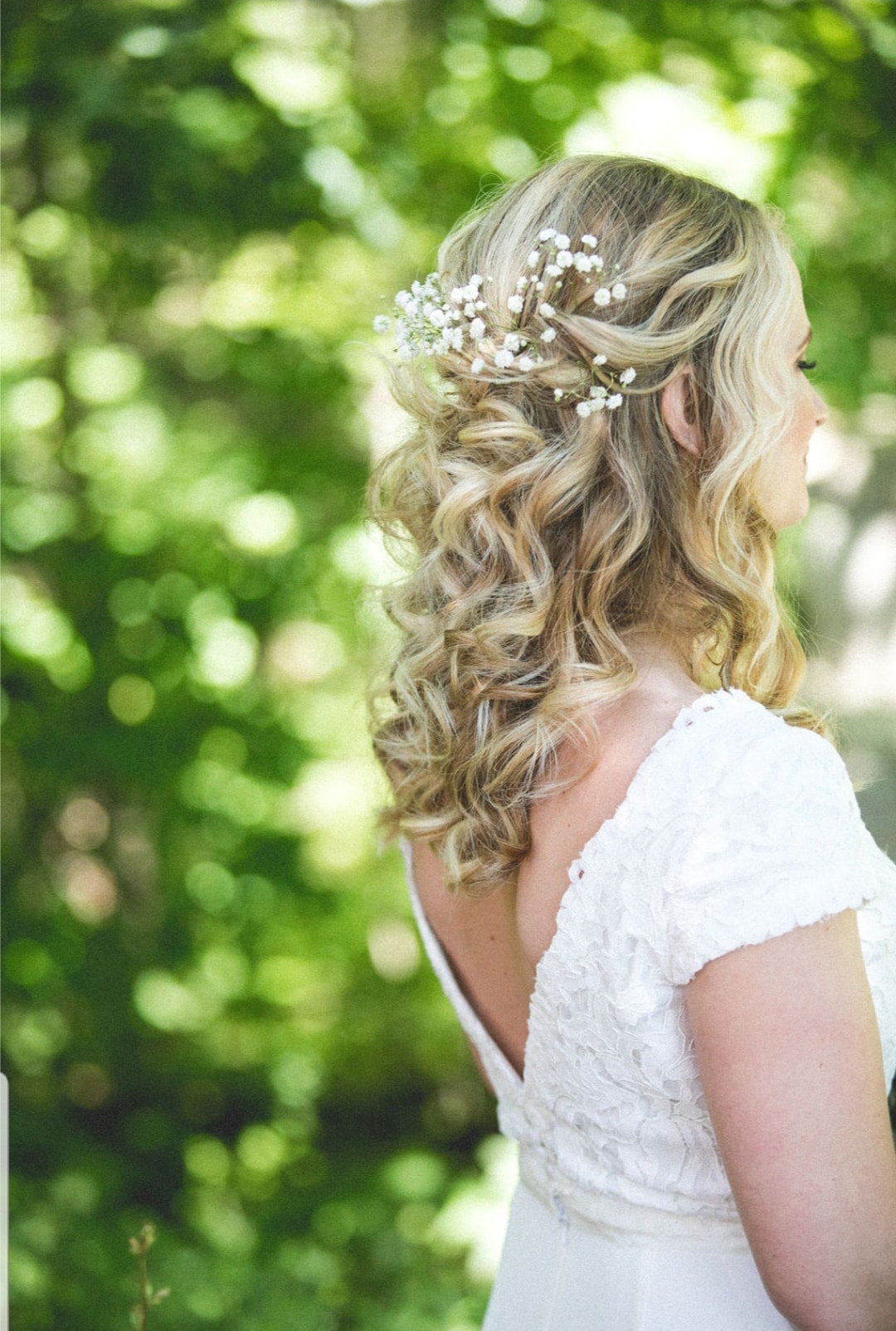 Side view of a woman with wavy blonde hair and a floral hair accessory, wearing a white dress outdoors.
