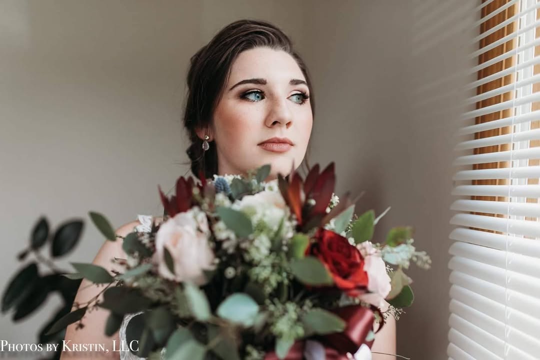 Woman with dark hair and makeup holding a bouquet of flowers near a window with blinds.