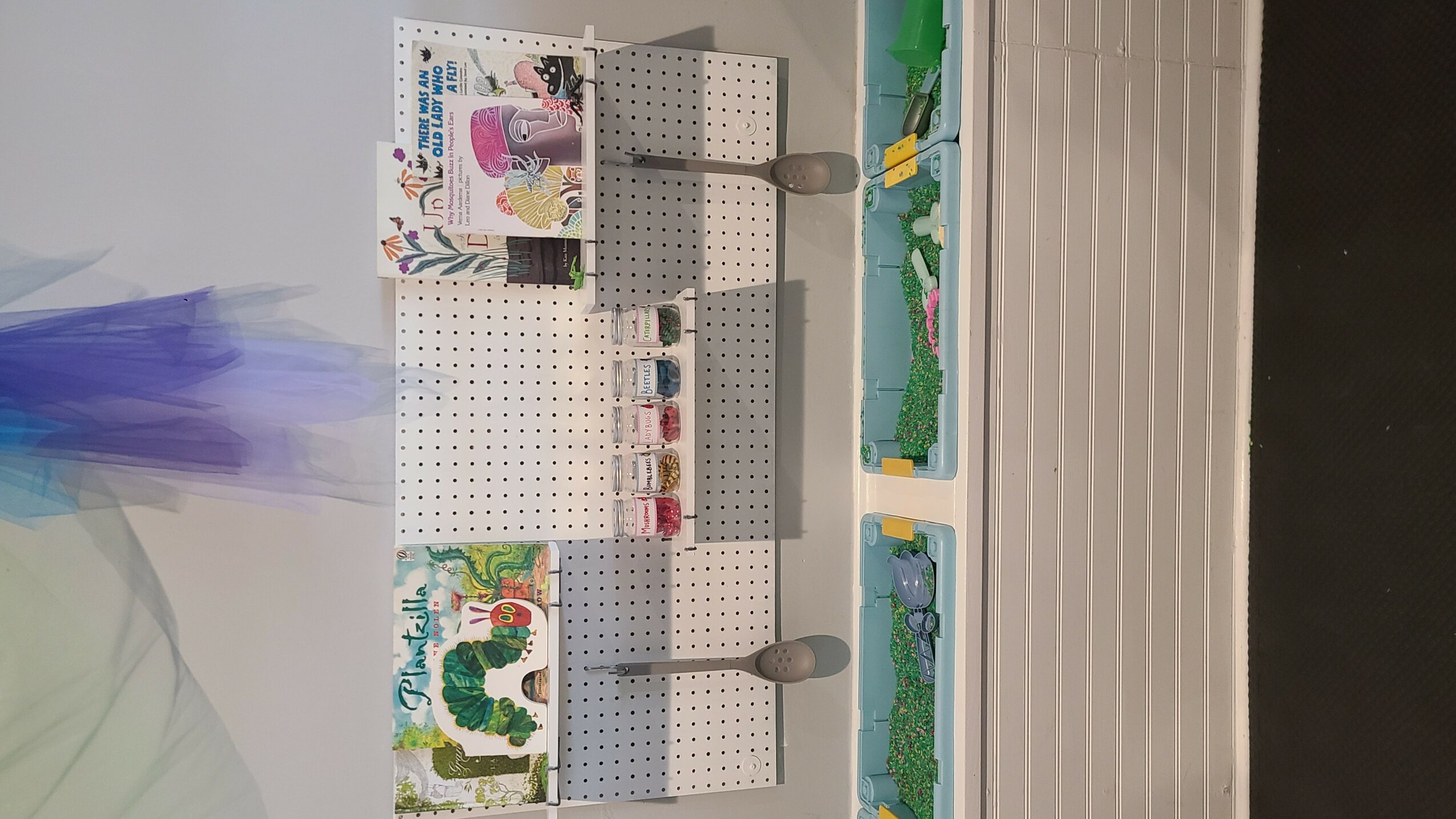 Display shelf with art supplies and paintings, viewed from the side, with a white wall and black floor.