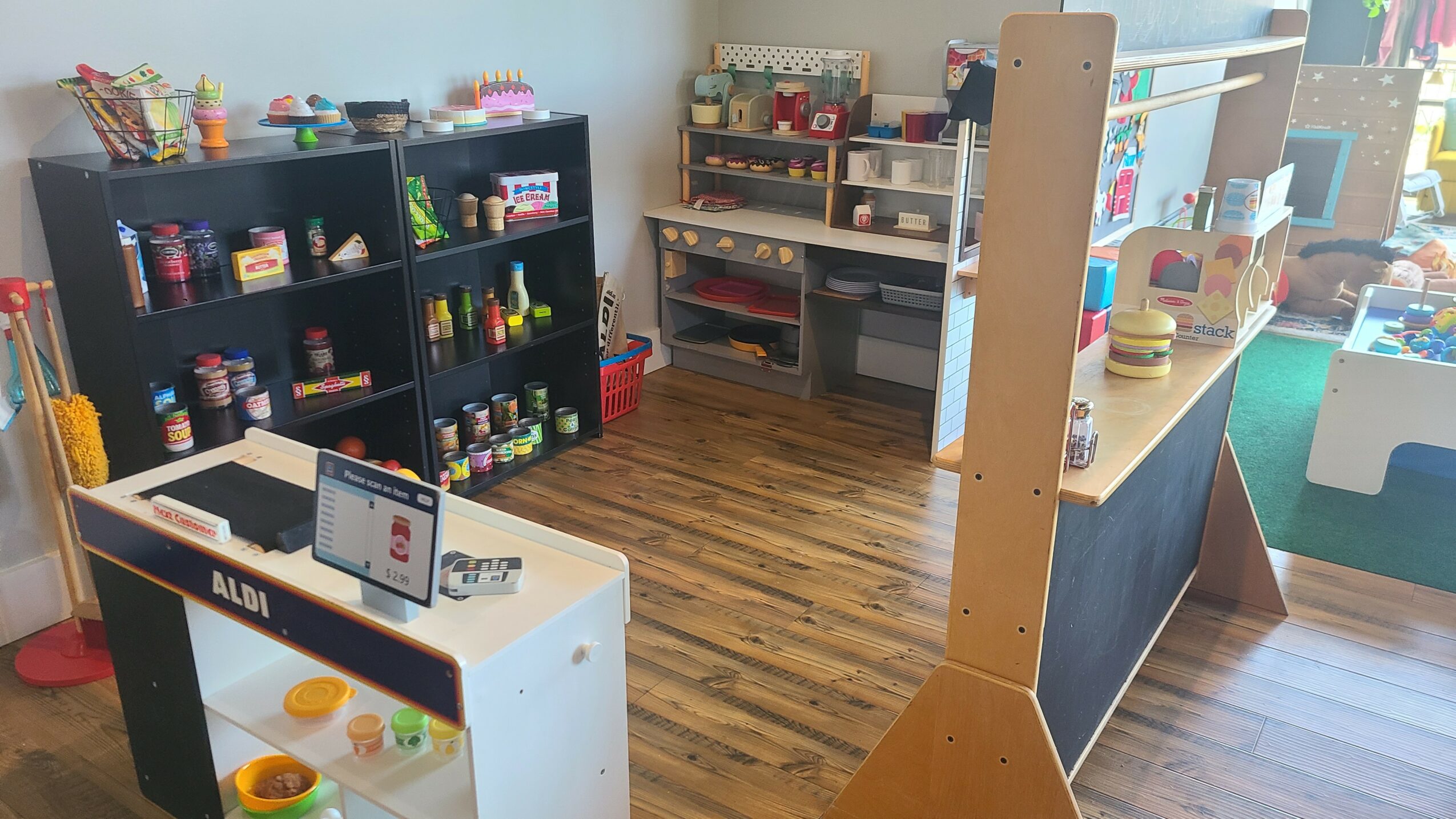 Indoor play area with shelves, tables, and toys, featuring wooden flooring and colorful play items.