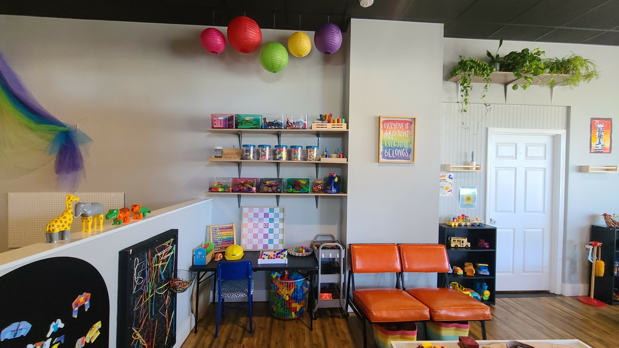 Colorful balloons hang above shelves with toys and books, orange seating, and a white door in a playroom.