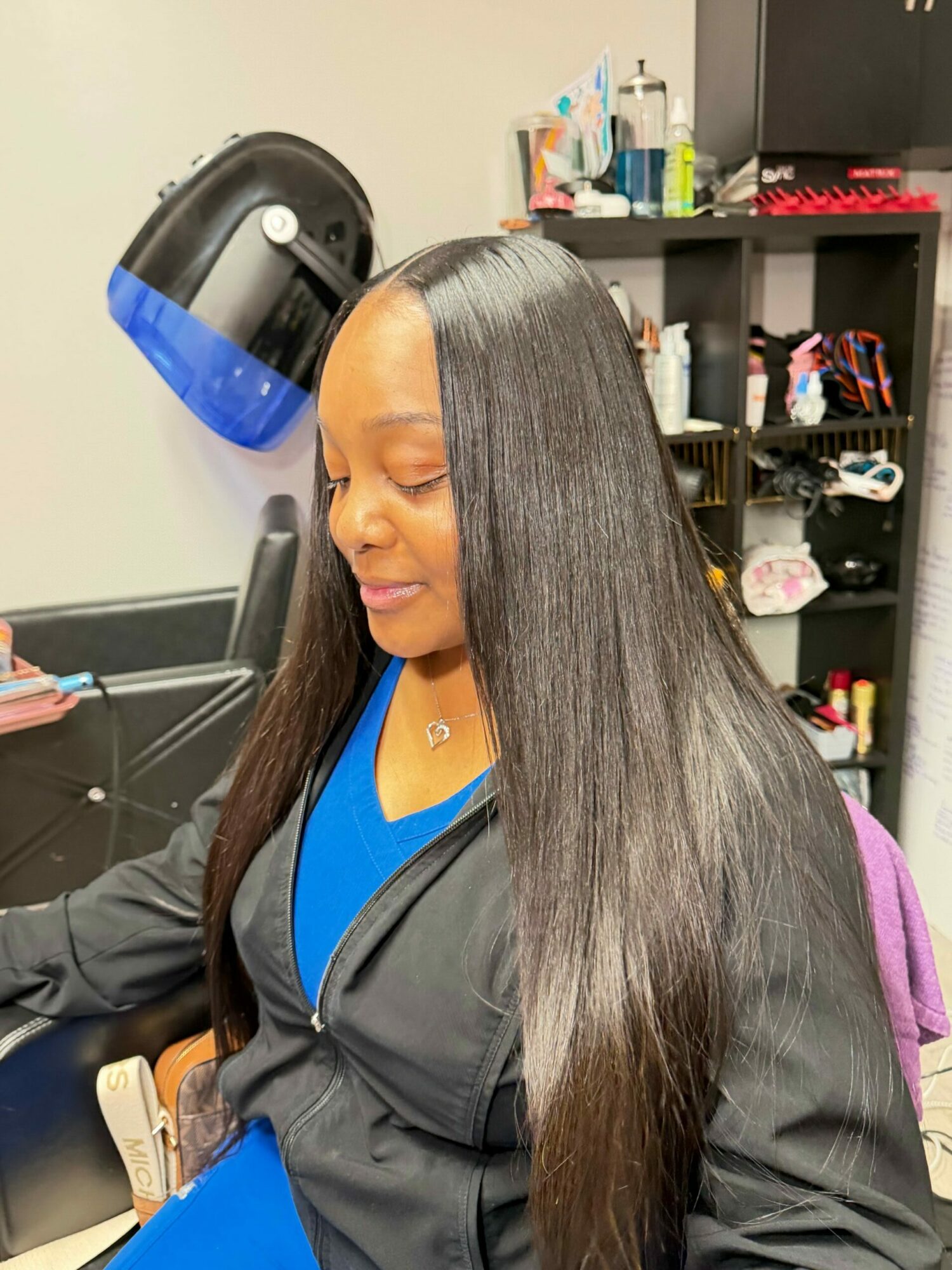 Woman with long straight hair wearing a black jacket and blue shirt, sitting in a room with shelves and various items.
