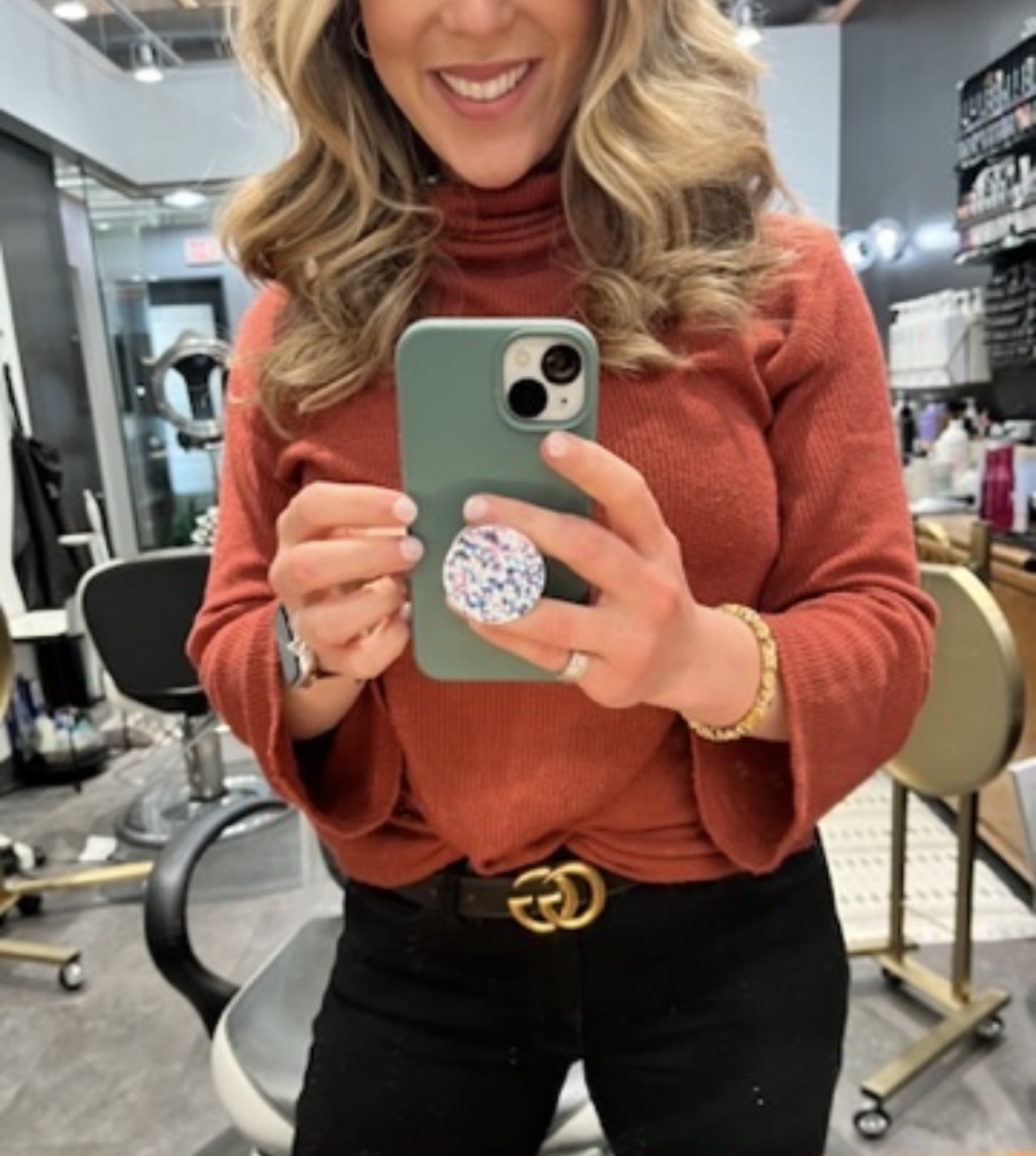 Woman taking a selfie in a salon, smiling, wearing a red top and black pants, holding a phone with a pop socket.