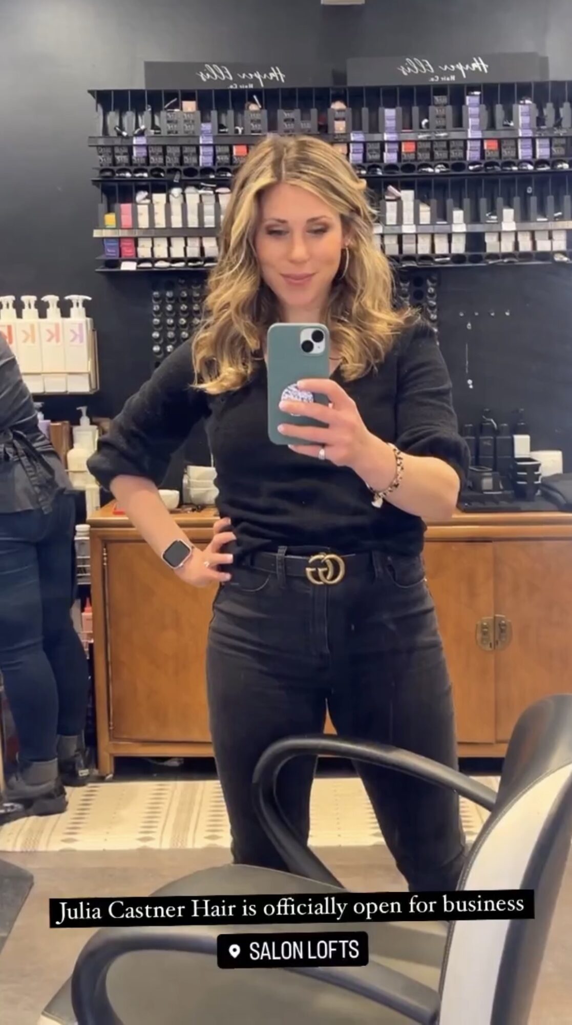 Woman with wavy blonde hair taking a selfie in a store, shelves with products behind her, wearing black outfit and belt.