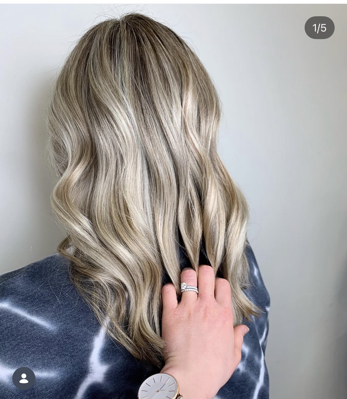 Person with wavy blonde hair, wearing a dark top, touching their hair with their hand, against a plain background.