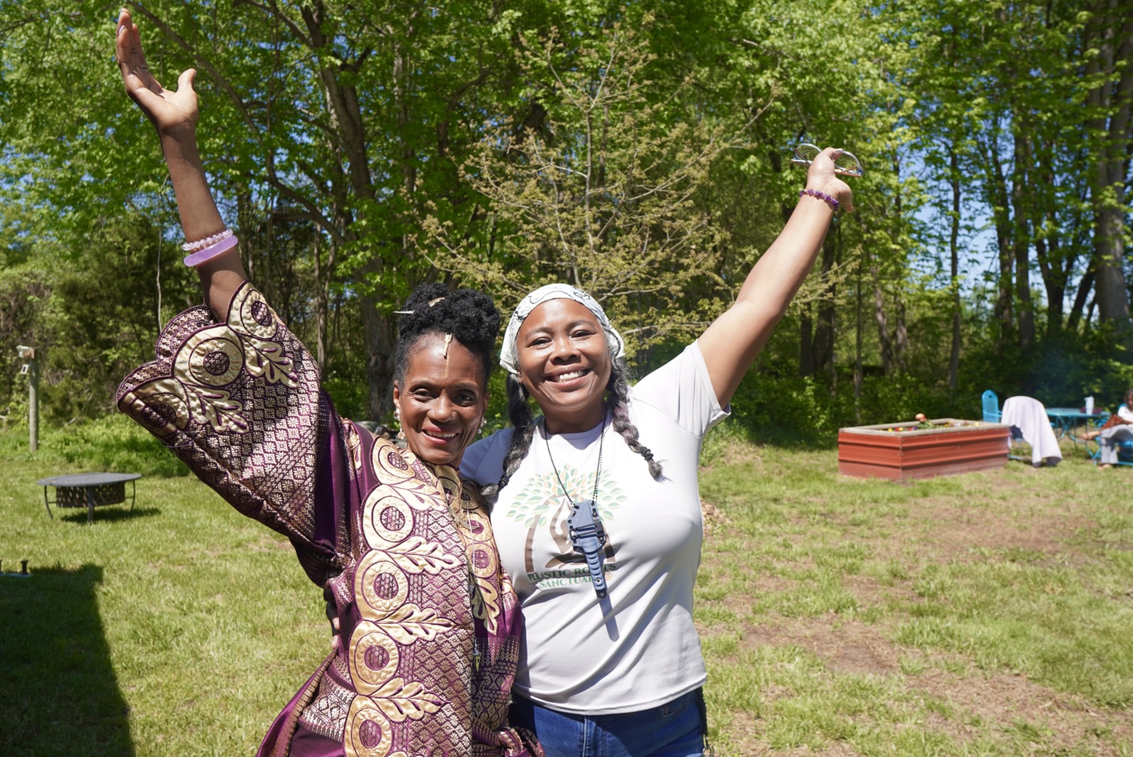 Two women smiling outdoors with arms raised, trees and grass in background.