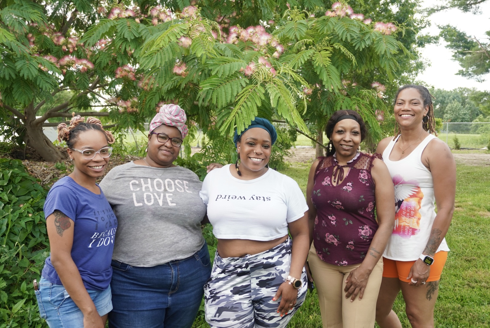 Five women standing outdoors under a flowering tree, smiling, with green grass and trees in background.