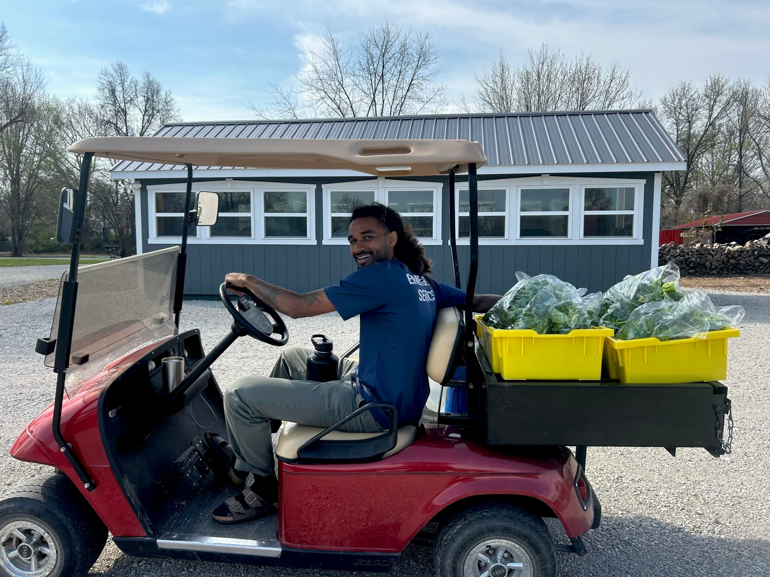 Person sitting in a red golf cart with yellow bins filled with green produce, parked outside a blue building with a metal roof.