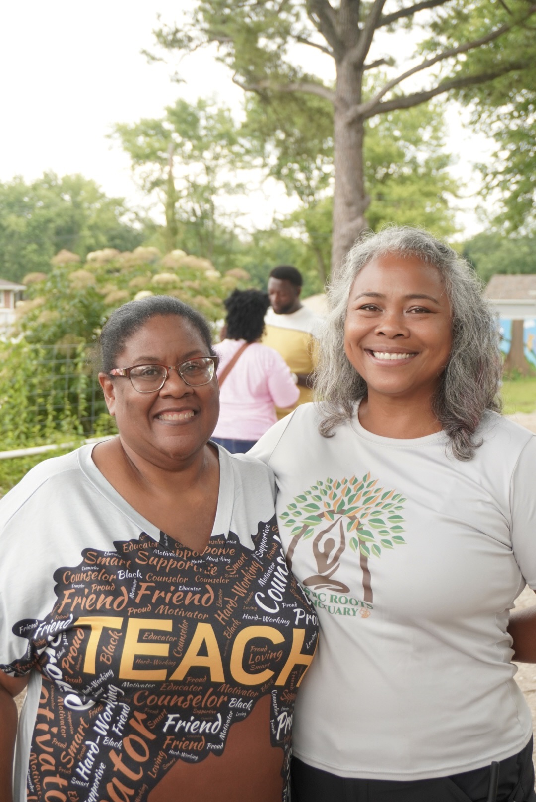 Two smiling women standing outdoors with trees and people in the background.