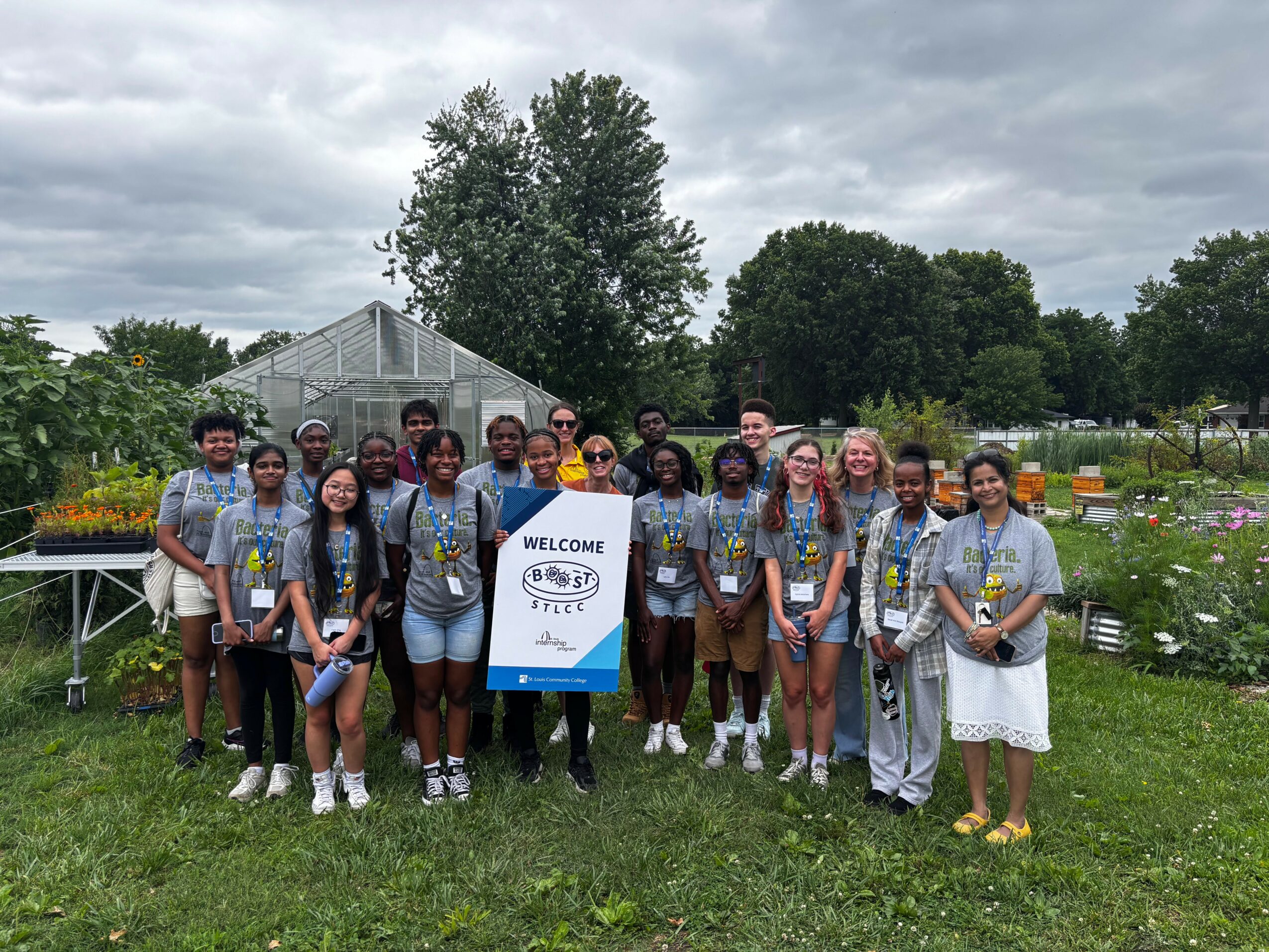 Group of people outdoors holding a welcome sign, standing on grass with trees and a greenhouse in background.