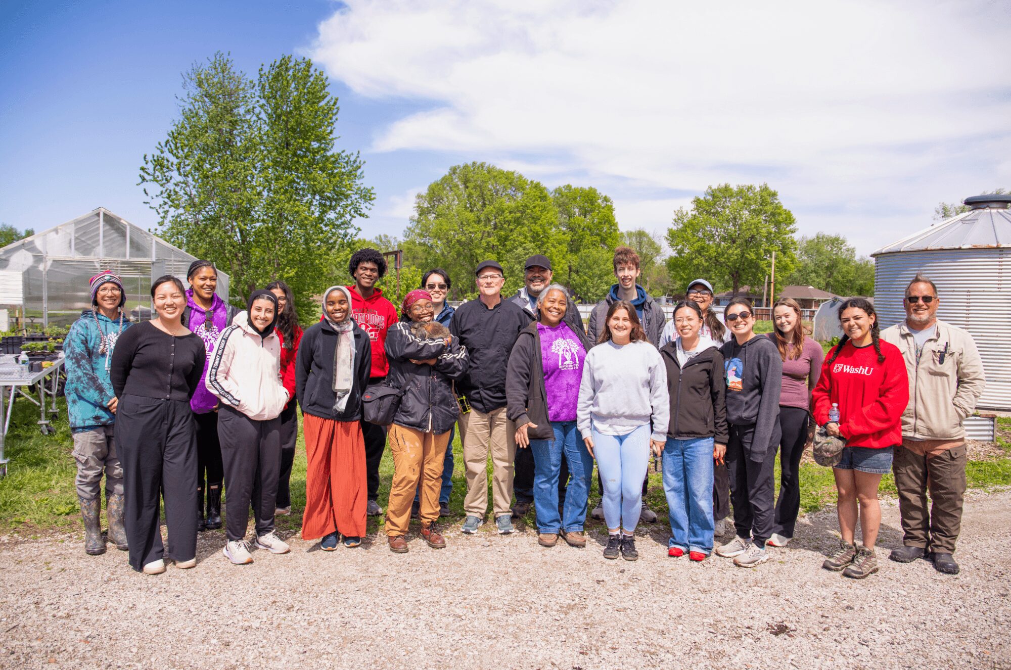 Group of people standing outdoors on a gravel path with trees and a cloudy sky in background.