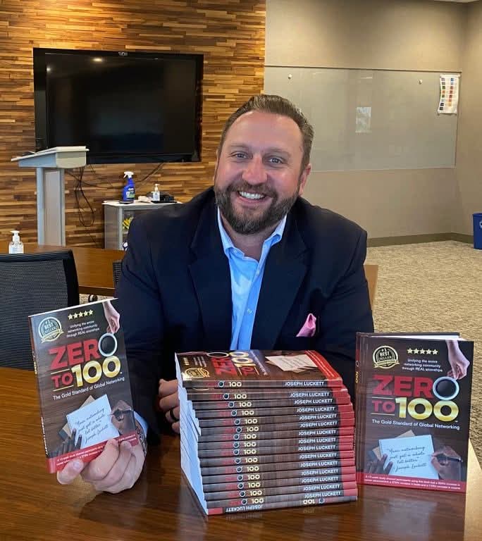 Man with a beard in a suit sitting at a table with stacks of books, smiling, in a room with a TV and wooden wall.