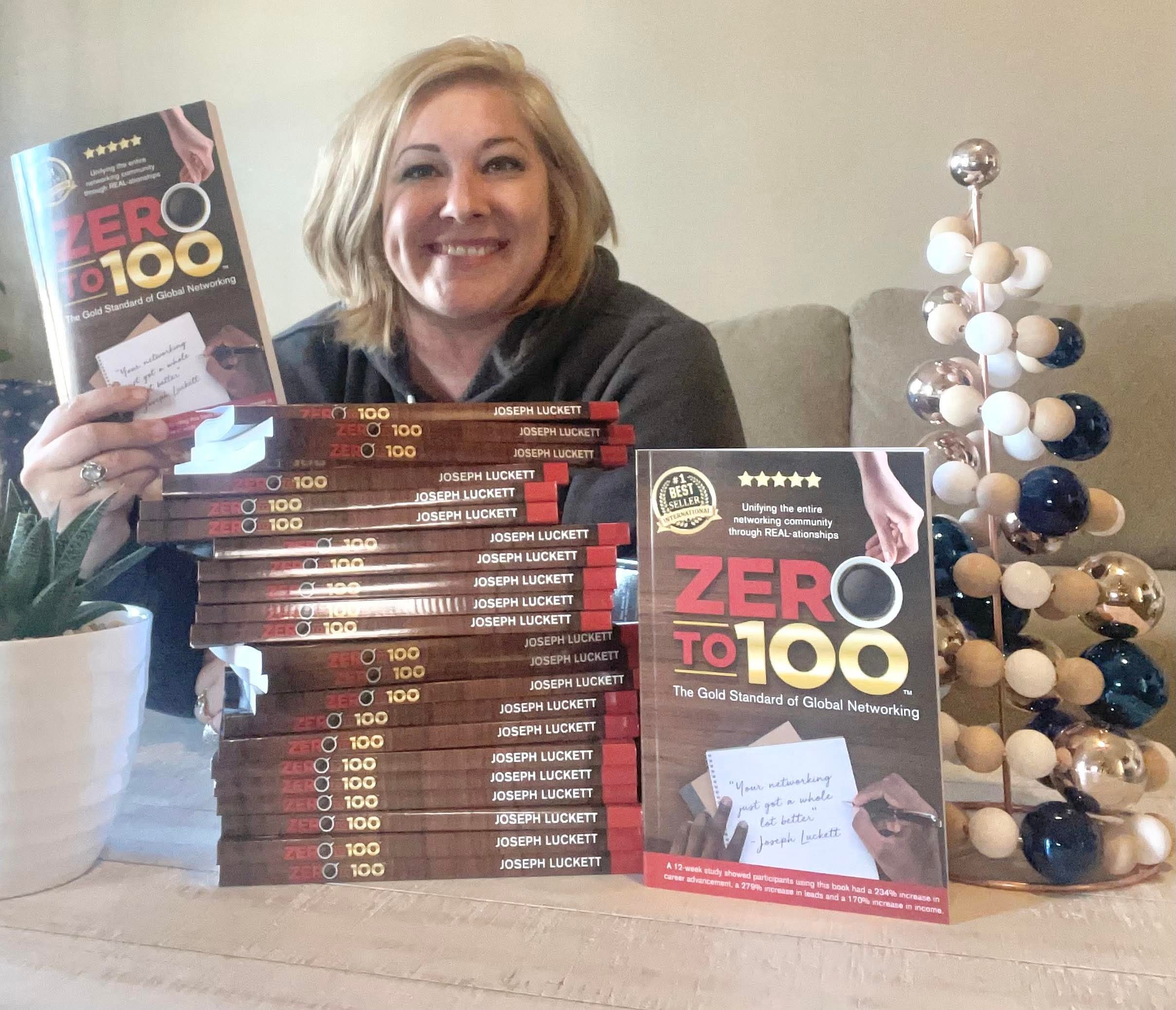 Woman smiling behind a stack of books titled 'Zero to 100' and a Christmas tree decoration on a table.