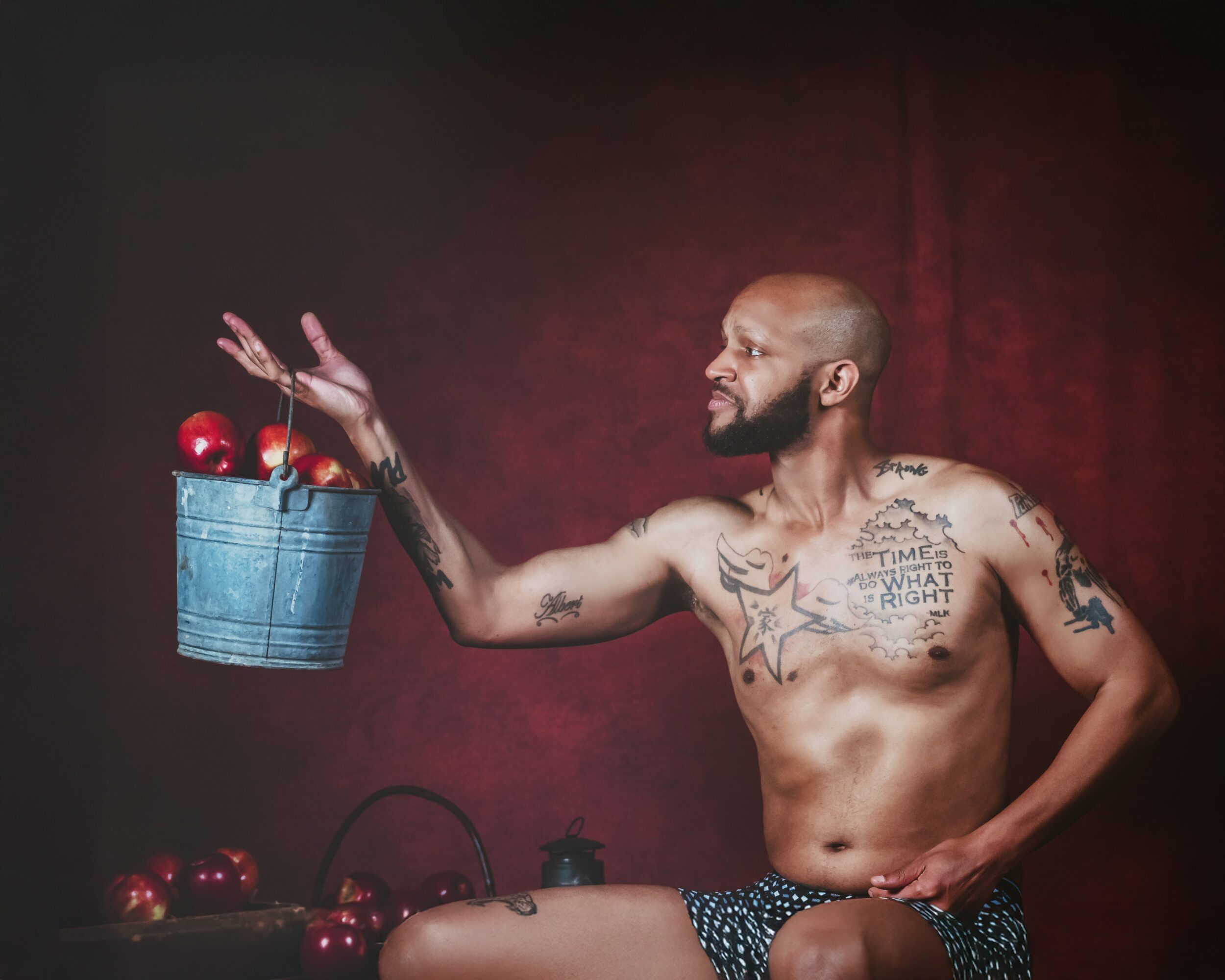 Bald man with tattoos sitting shirtless, holding a bucket of red apples, against a dark red background.