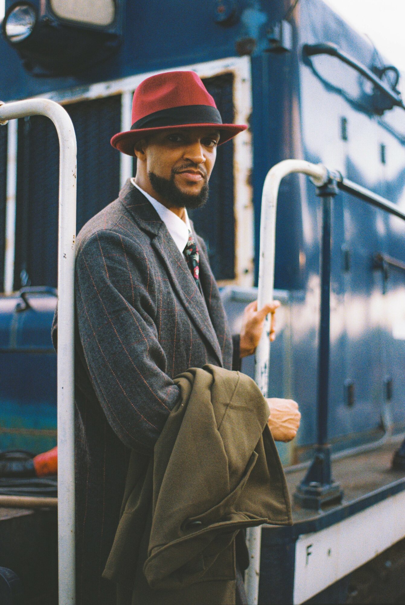 Man in a gray suit and red hat standing on a train platform holding a coat, with a blue train in the background.