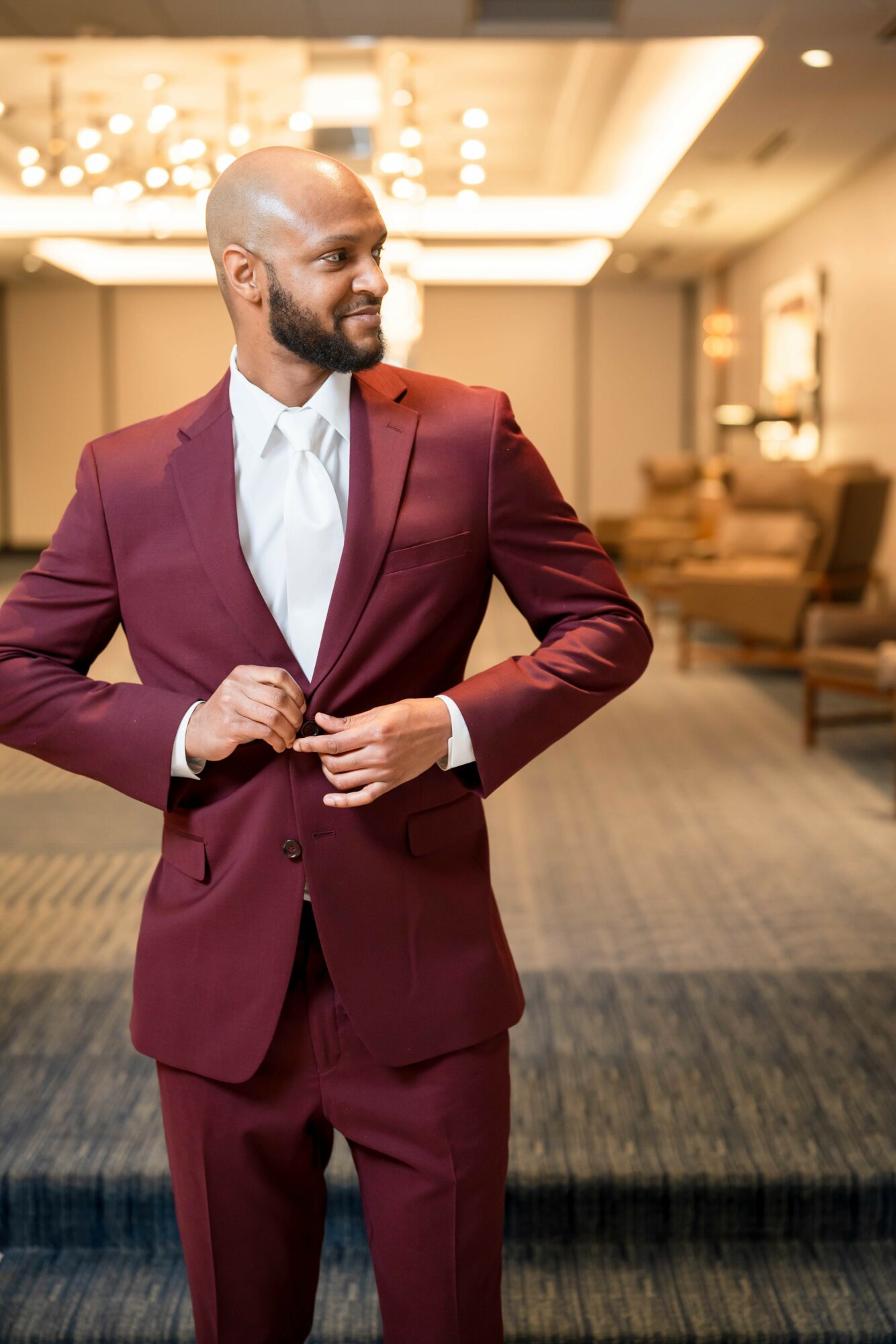 Man in burgundy suit adjusting his jacket in a well-lit, elegant room with chairs and a chandelier.