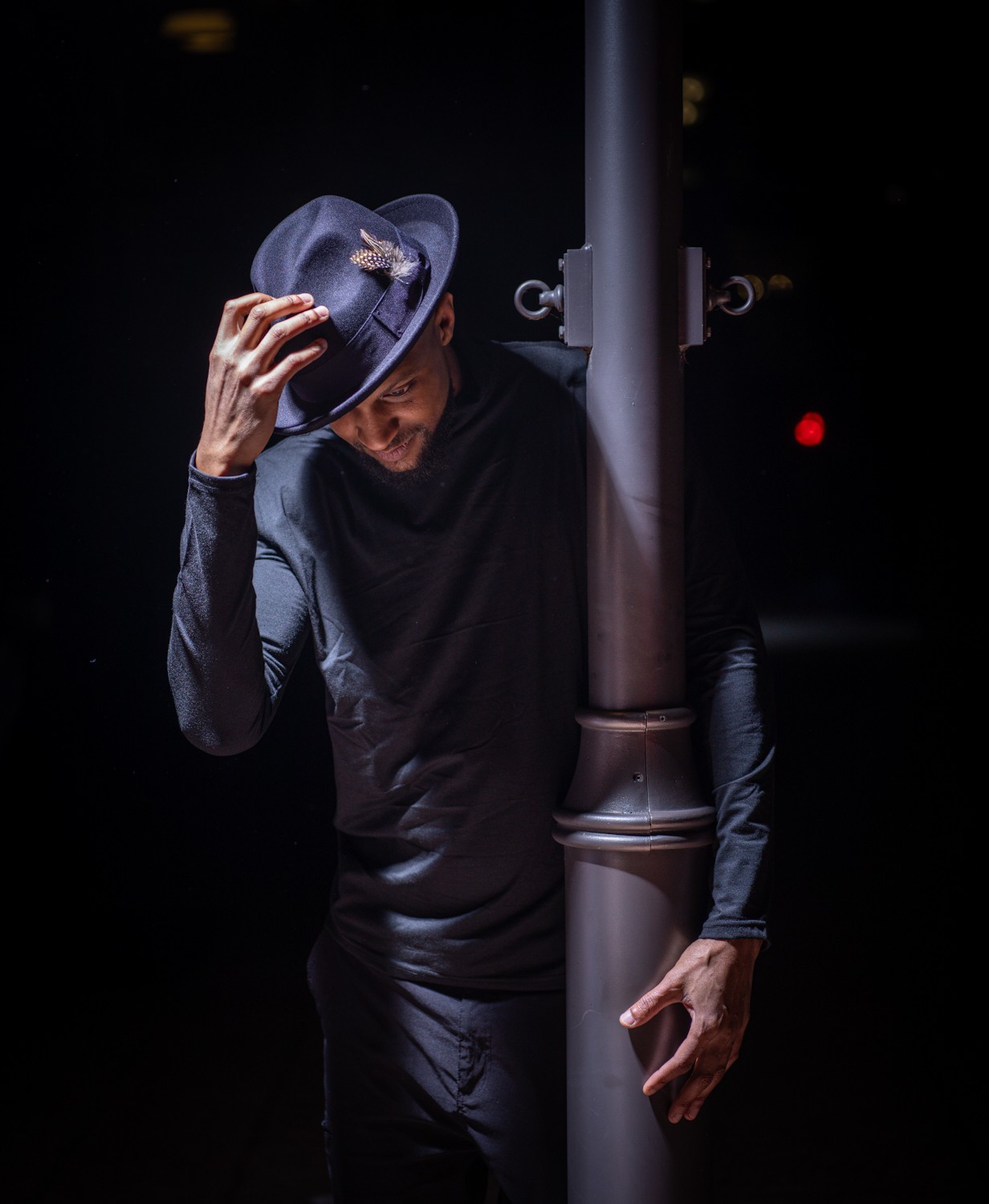 Person wearing a hat and dark clothing, leaning against a streetlamp in a dark setting.