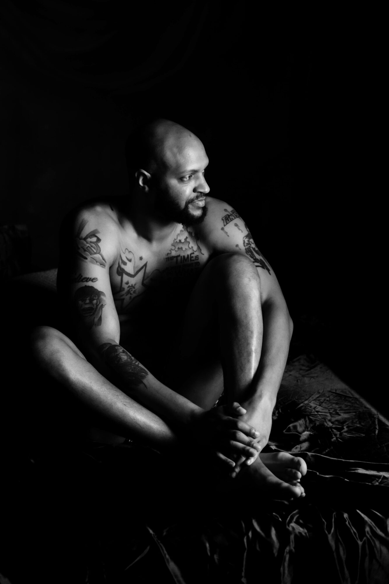 Man with tattoos sitting on a bed, looking to the side, in a dark room with minimal lighting.