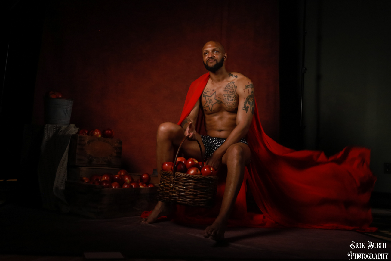 Man with tattoos sitting on a stool, surrounded by apples and a basket, draped in a red cloth, dark background.