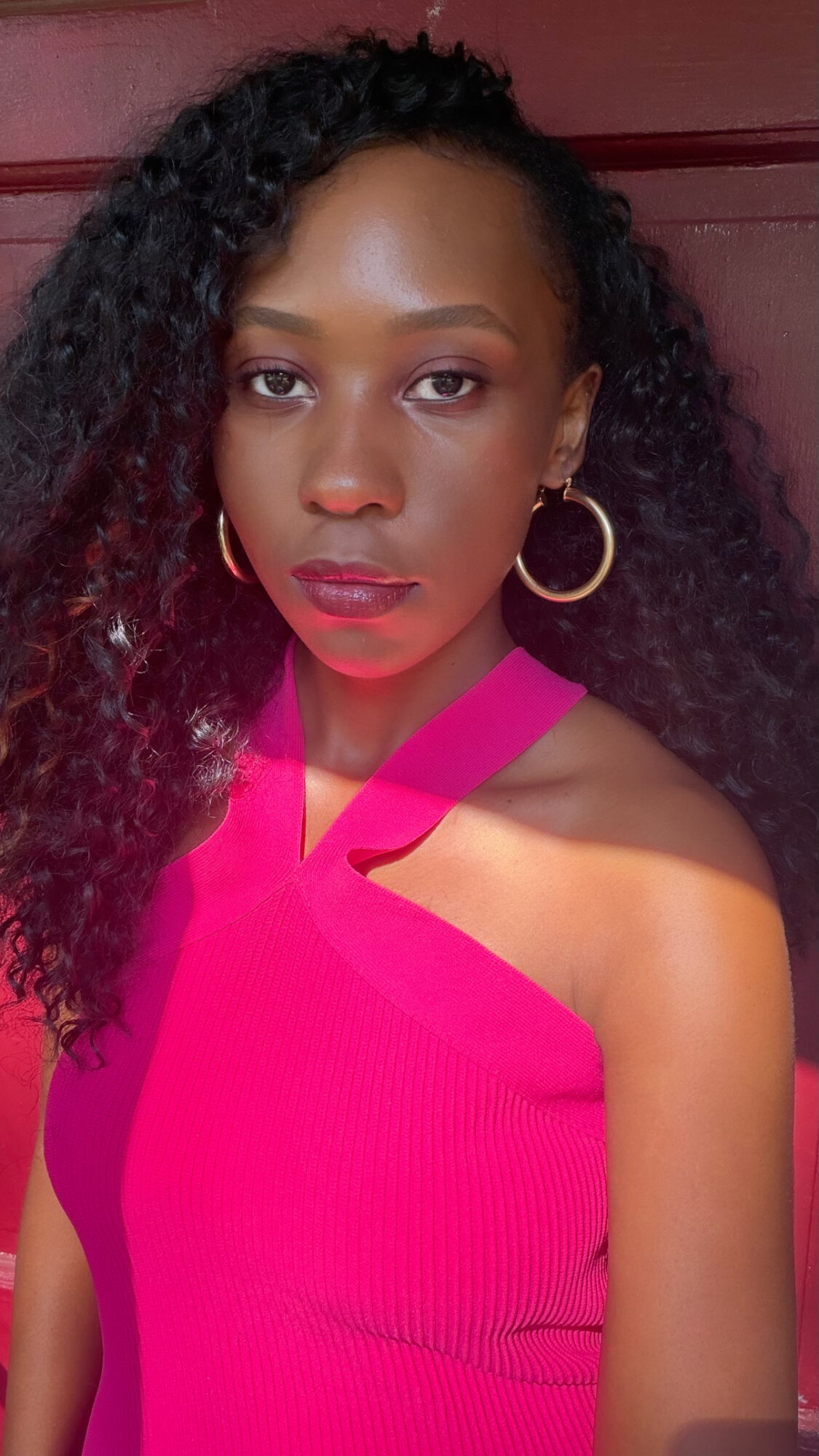Young woman with curly hair wearing a bright pink top and large hoop earrings, looking at the camera.