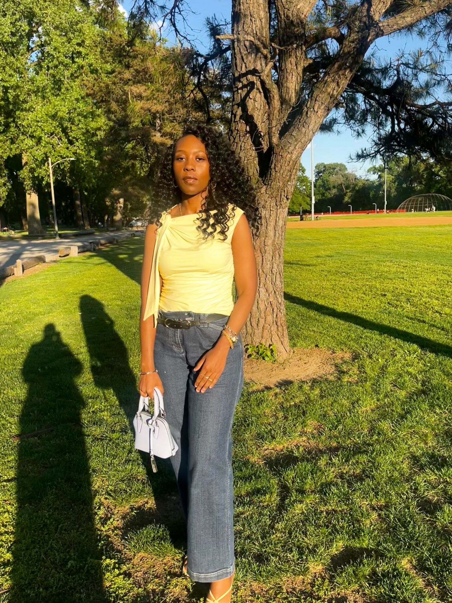 Woman standing outdoors near a tree, wearing a yellow top and jeans, holding a white bag, with sunlight and green grass.