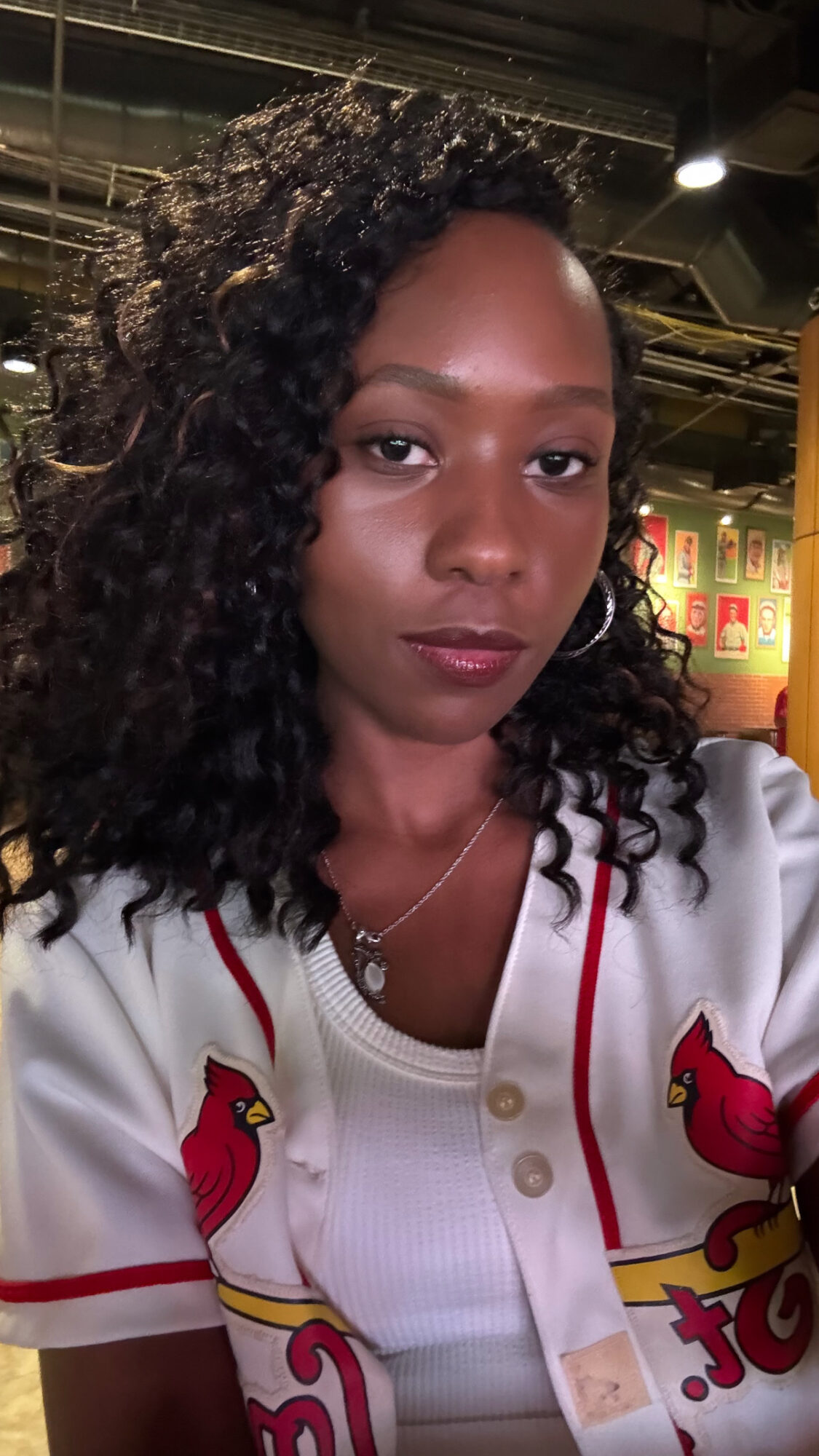 Woman with curly hair wearing a white sports jersey with red and yellow logos, indoors with ceiling lights and decorations