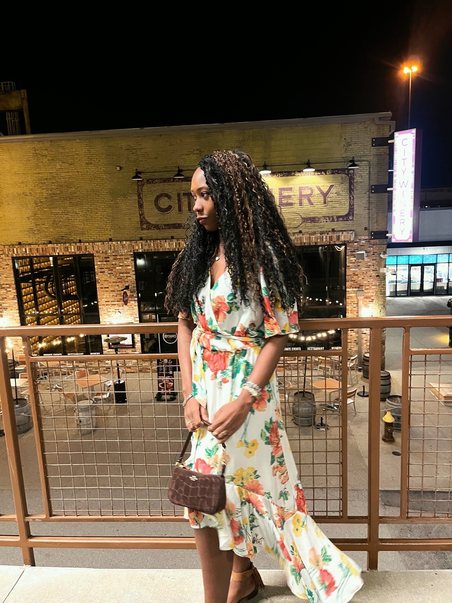 Woman with curly hair in a floral dress holding a purse, standing outdoors at night near a railing.