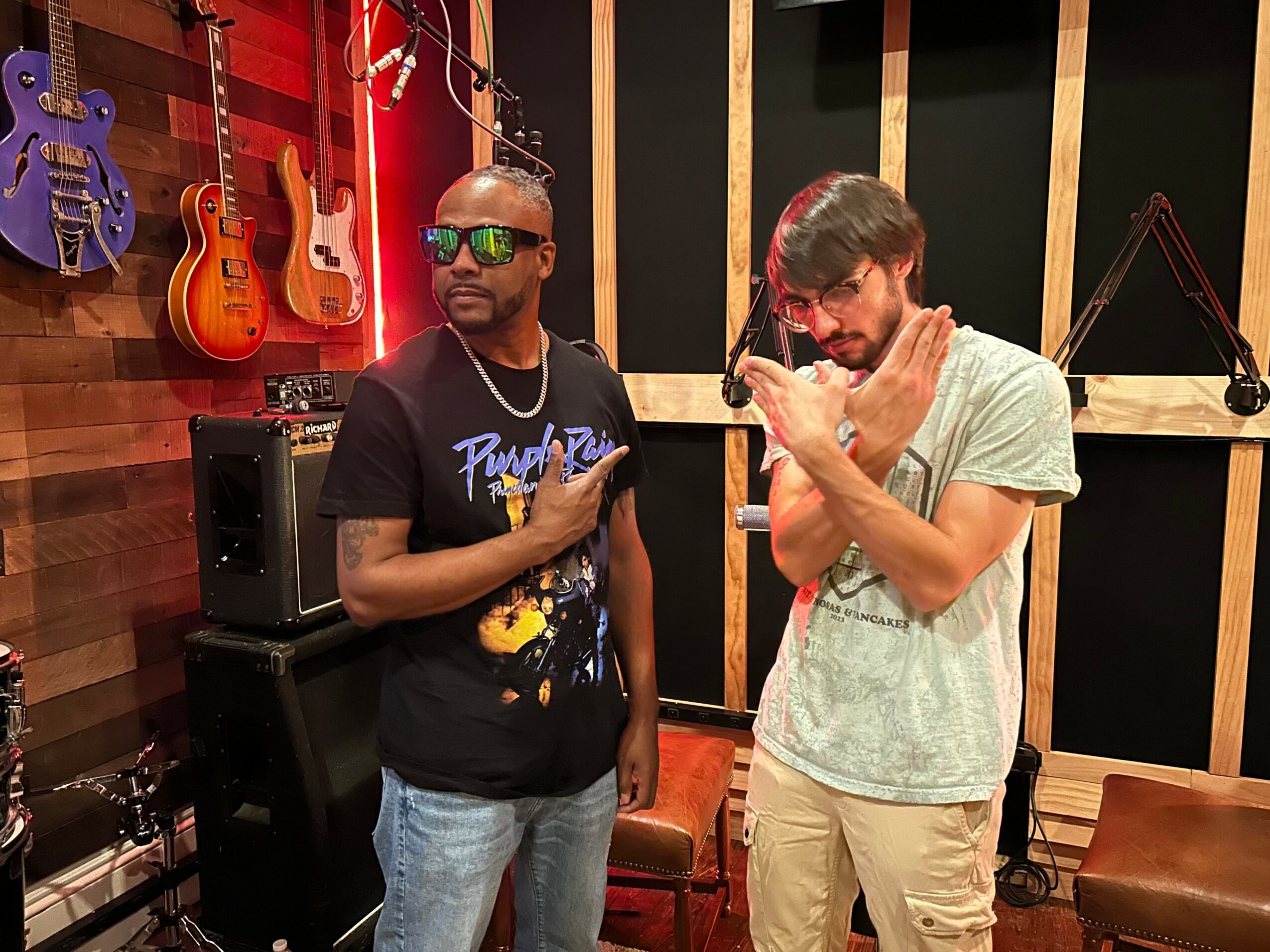 Two men in a music studio with guitars hanging on the wall, one wearing sunglasses and the other with glasses, both looking at their phones.