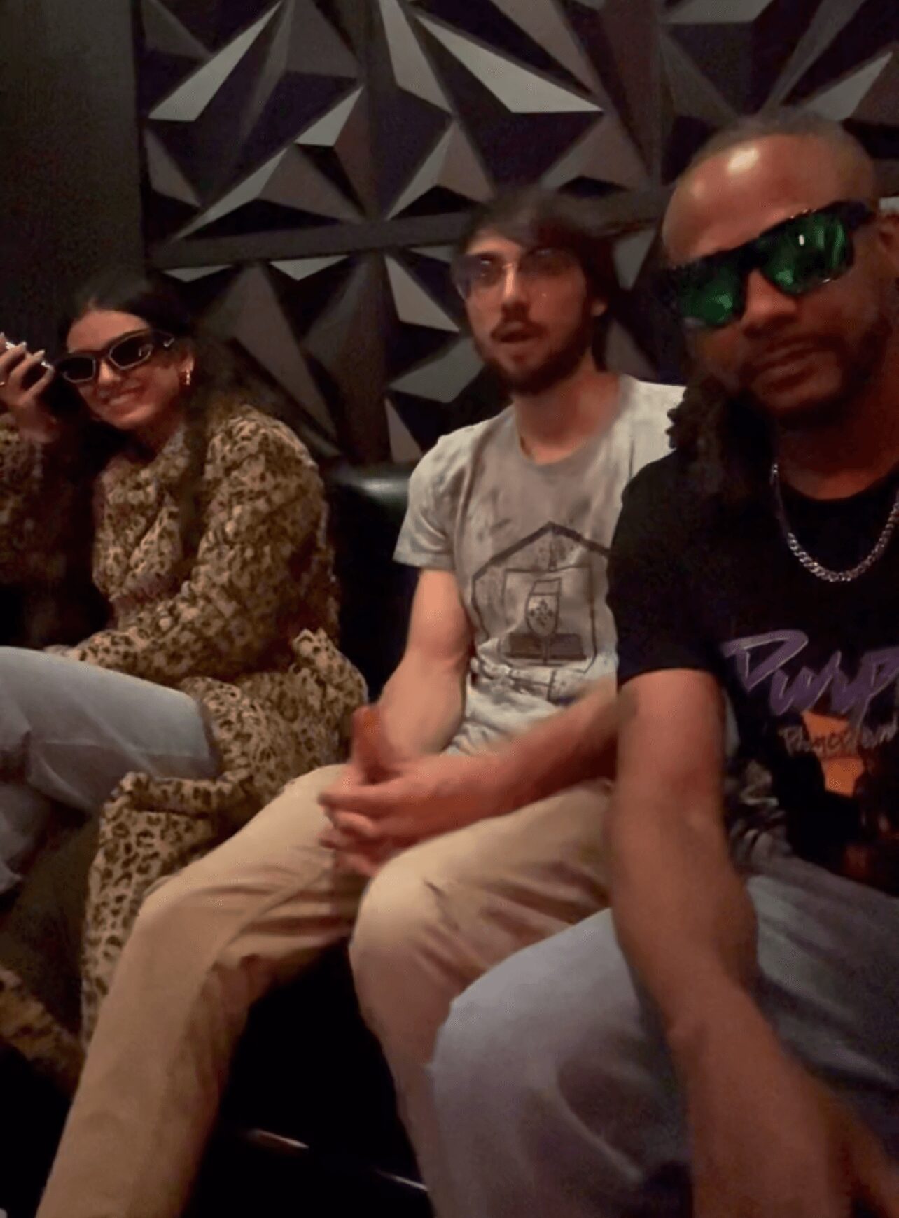 Three people sitting together indoors, two wearing sunglasses, one with a beard, in front of a geometric wall pattern.