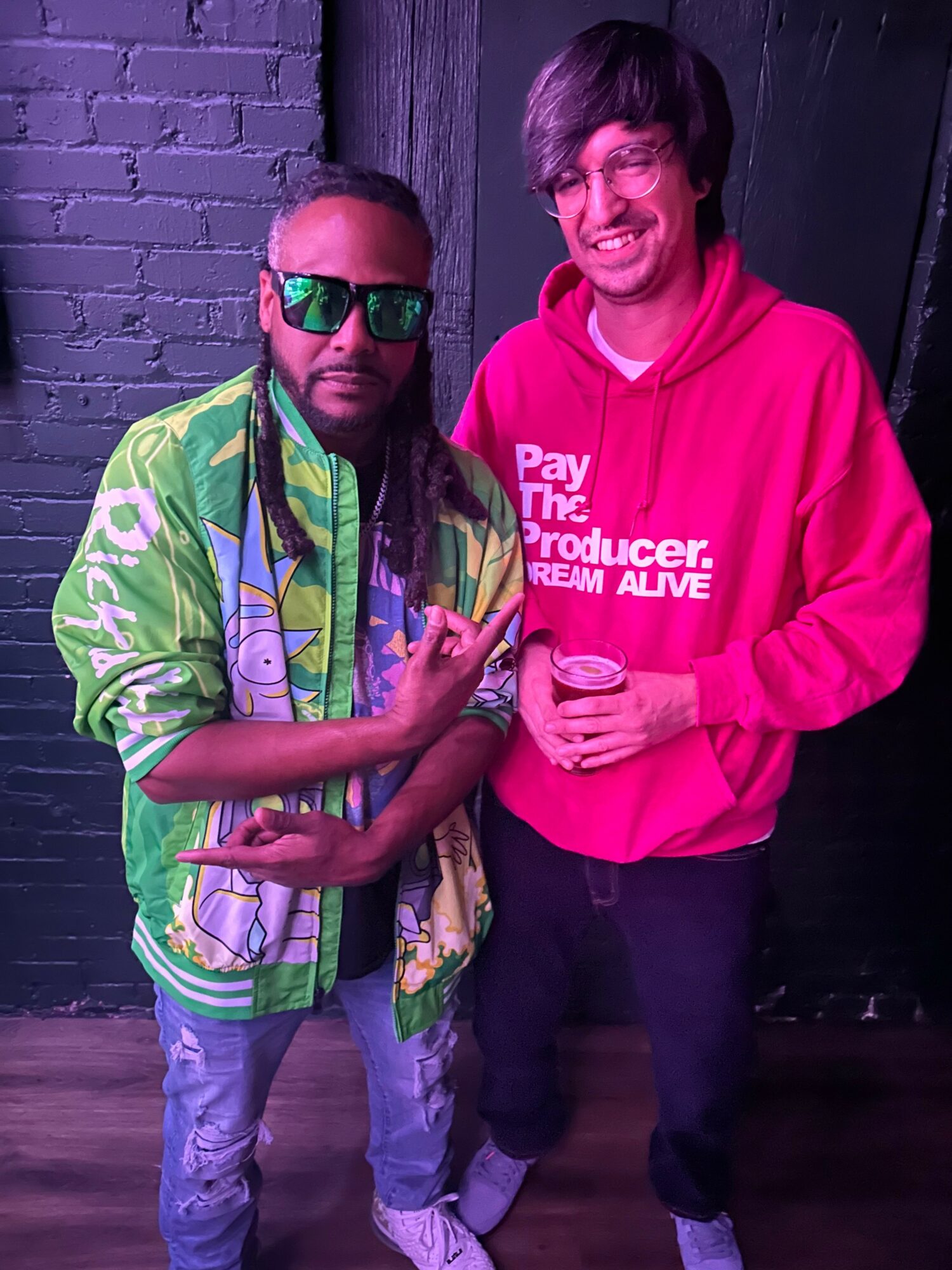 Two people standing together, one wearing sunglasses and a colorful jacket, the other in a red hoodie holding a cup.