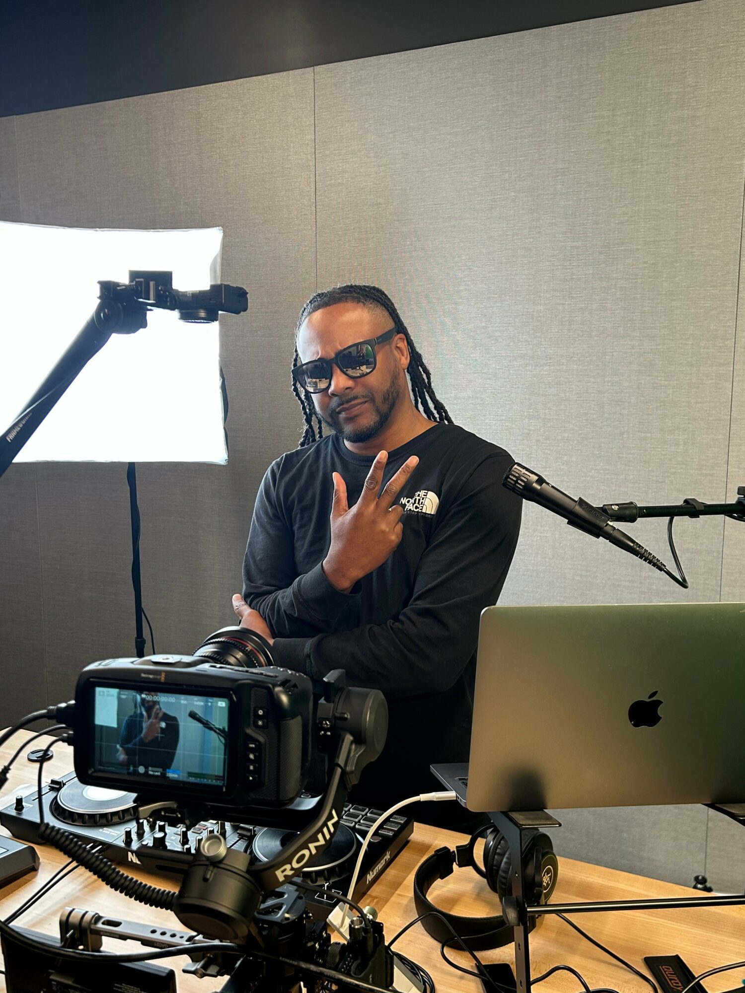 Person with sunglasses and braided hair making a hand gesture, sitting at a desk with camera and laptop, in a studio.