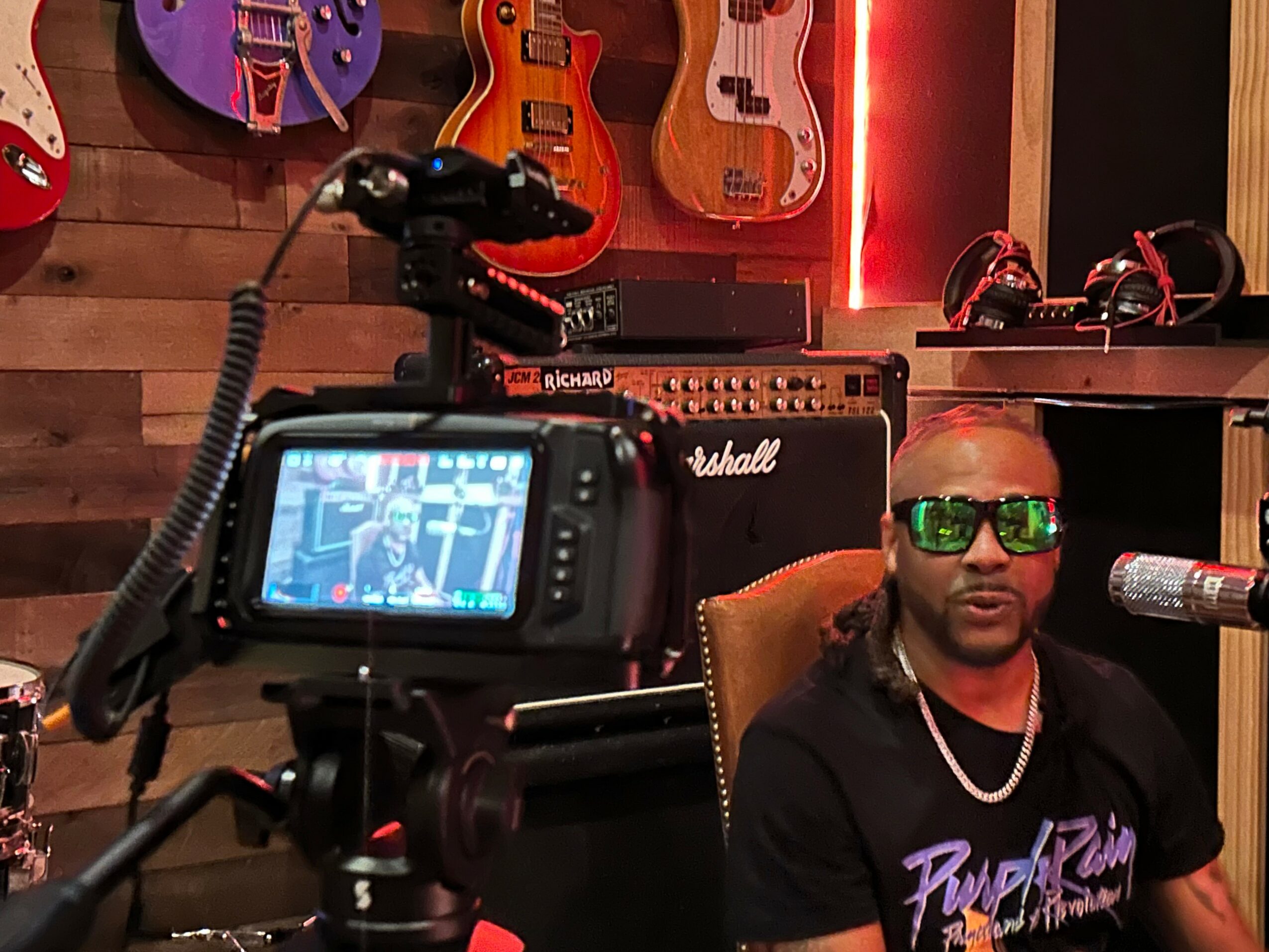 Man with sunglasses and a chain speaking into a microphone, with guitars and amplifiers in the background.