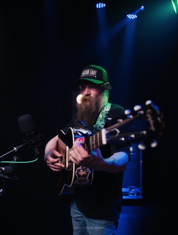 Bearded man wearing a cap playing an acoustic guitar on stage with colorful lights.