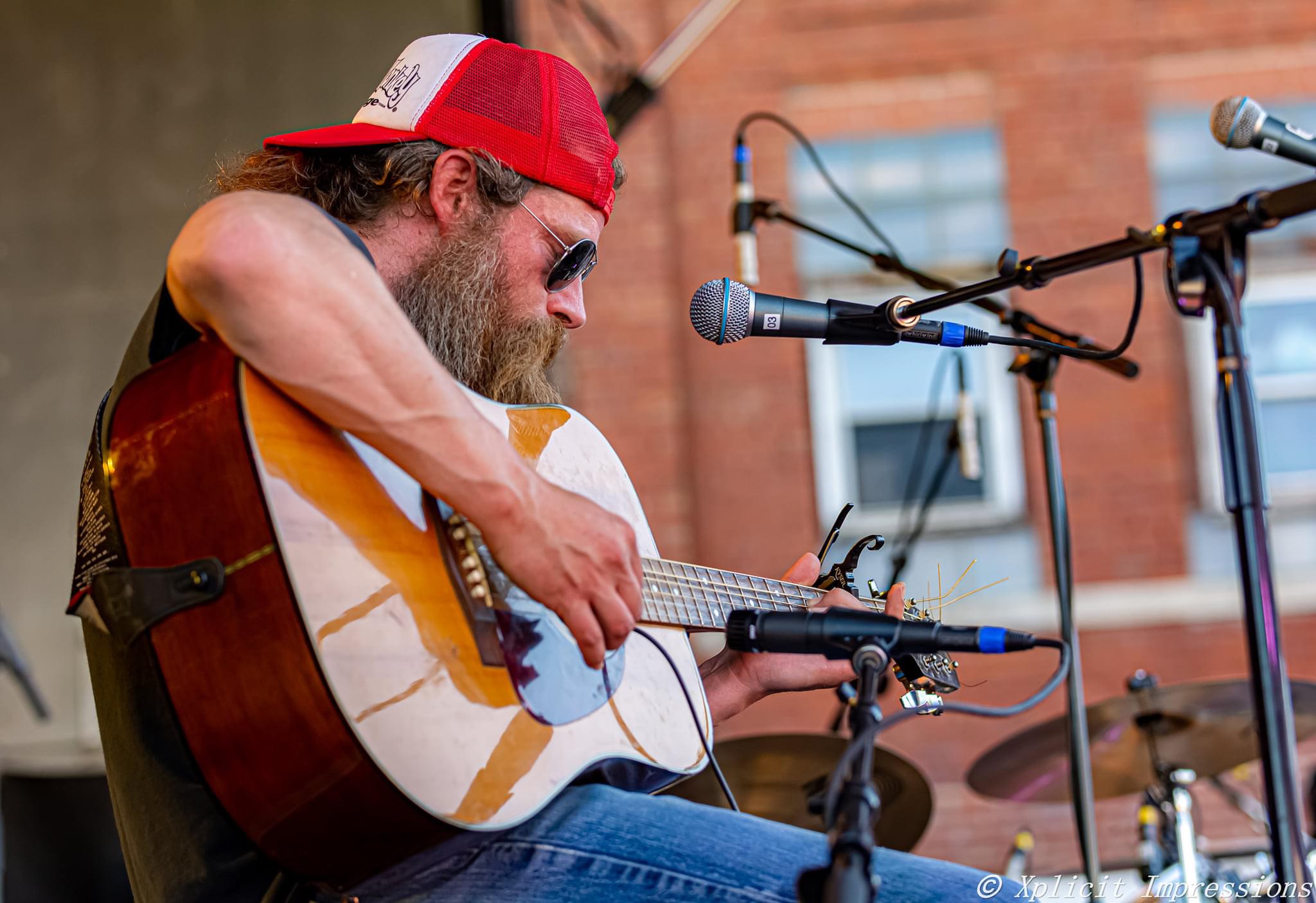 Man playing guitar and singing into microphone outdoors, brick building in background.