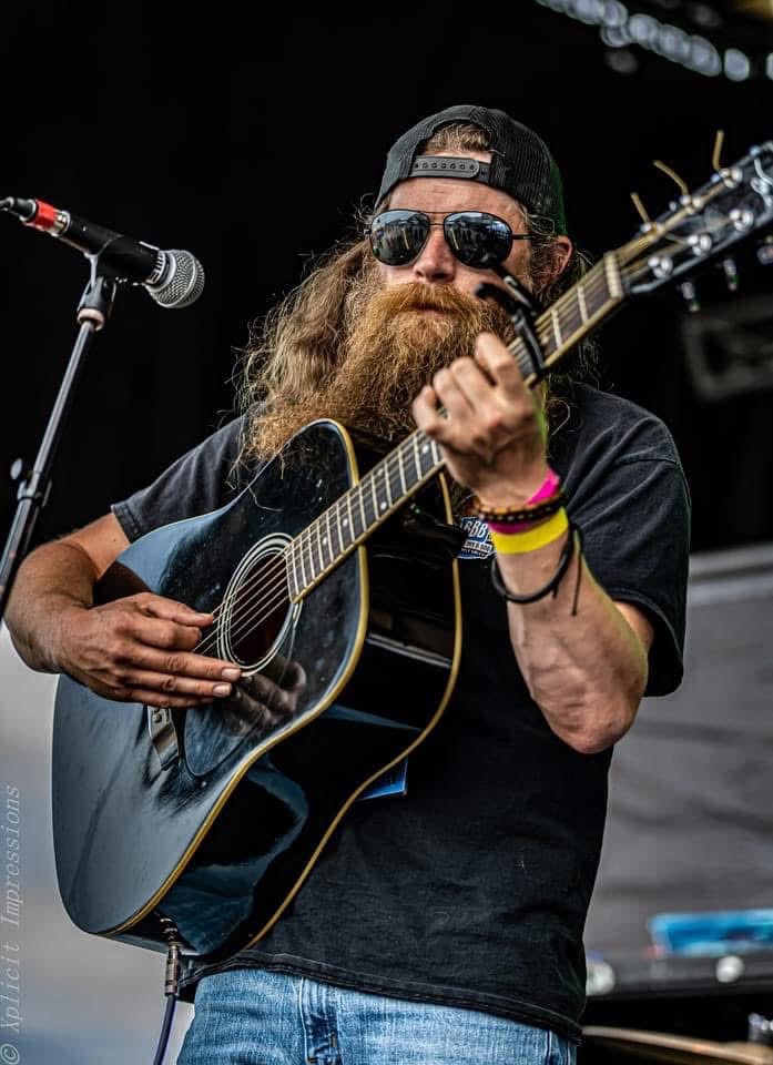 Man with long hair, beard, sunglasses, and a cap playing an acoustic guitar on stage.