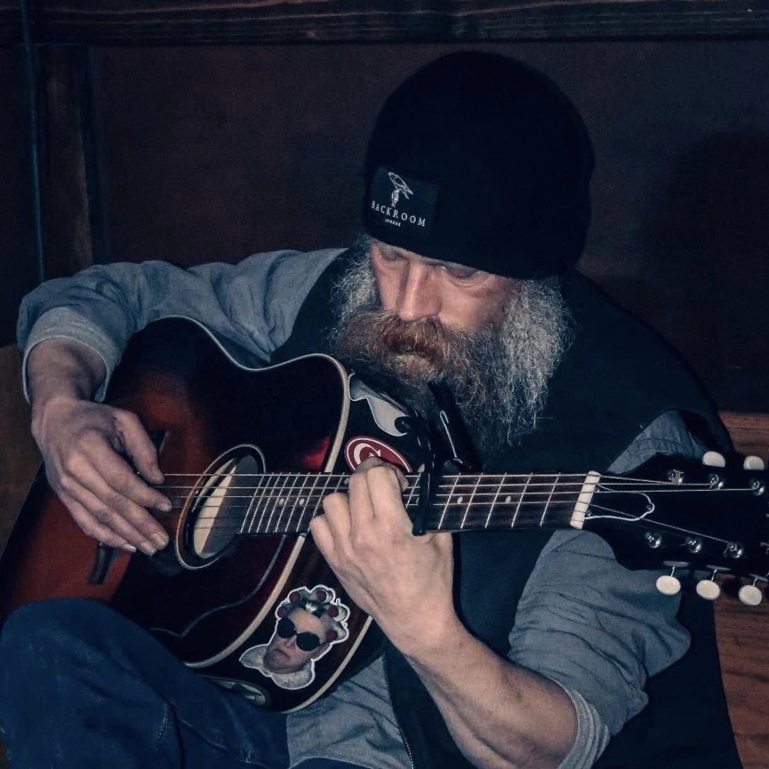 Man with a beard wearing a black beanie playing an acoustic guitar, sitting indoors in a dimly lit setting.