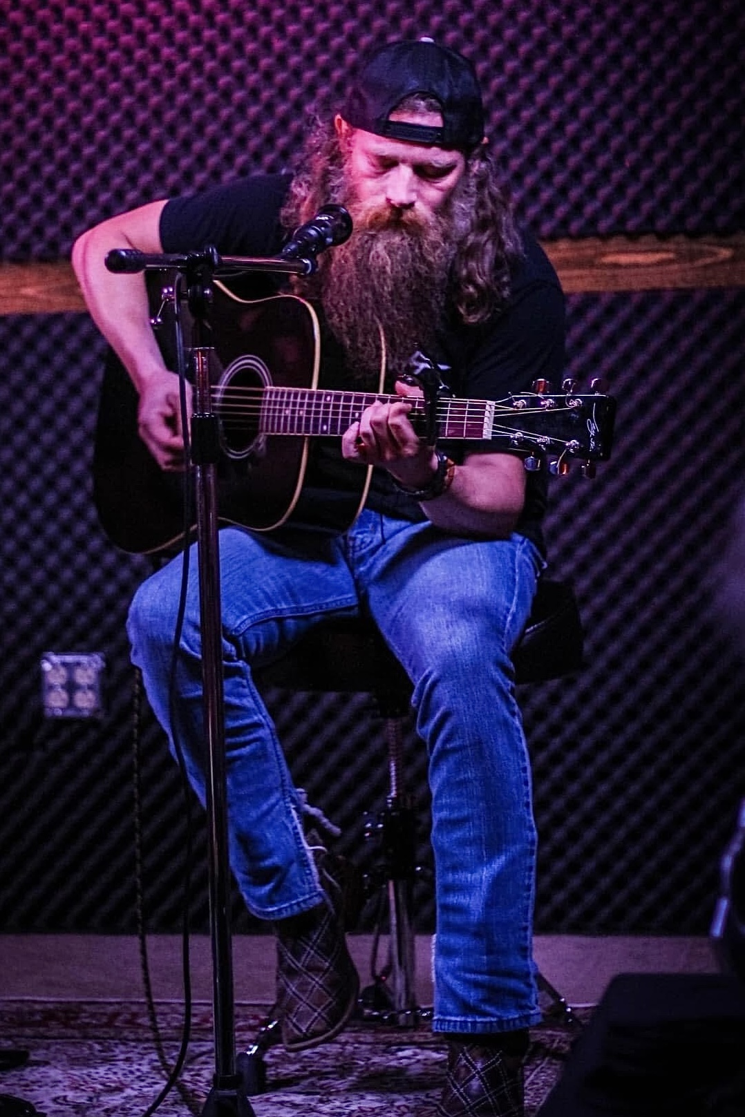 Bearded man wearing a cap playing an acoustic guitar on stage with a microphone, seated on a stool.