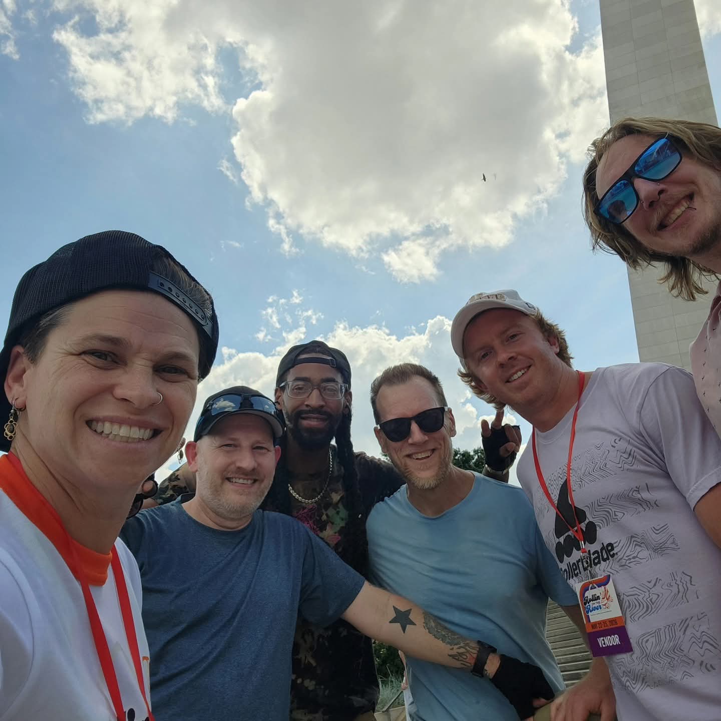 Group of six people smiling outdoors with a partly cloudy sky, some wearing sunglasses and lanyards.