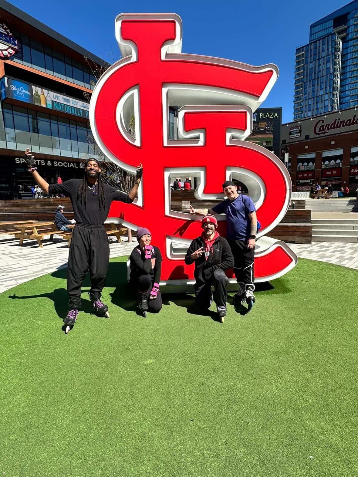 Four people pose in front of a large red and white logo on a green lawn, with modern buildings in background.
