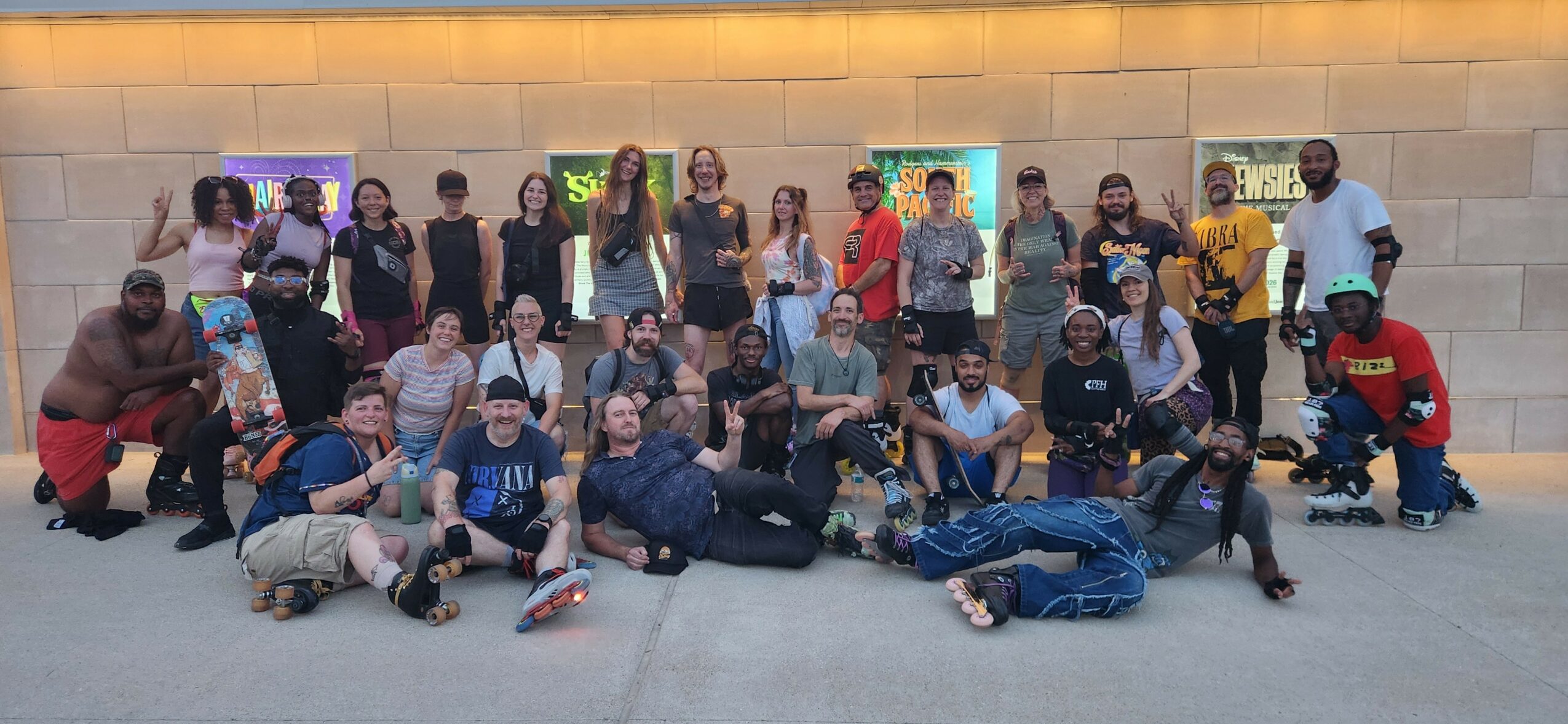 Group of people posing outdoors in front of a wall with colorful artwork, some sitting and some standing.