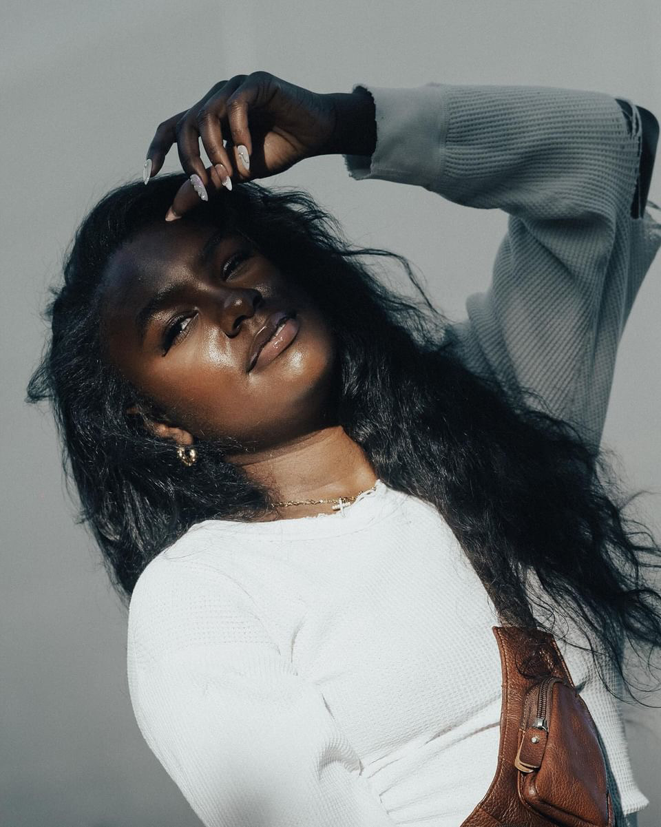 Woman with long dark hair, wearing a white top and gray jacket, poses with hand on head, looking at camera.