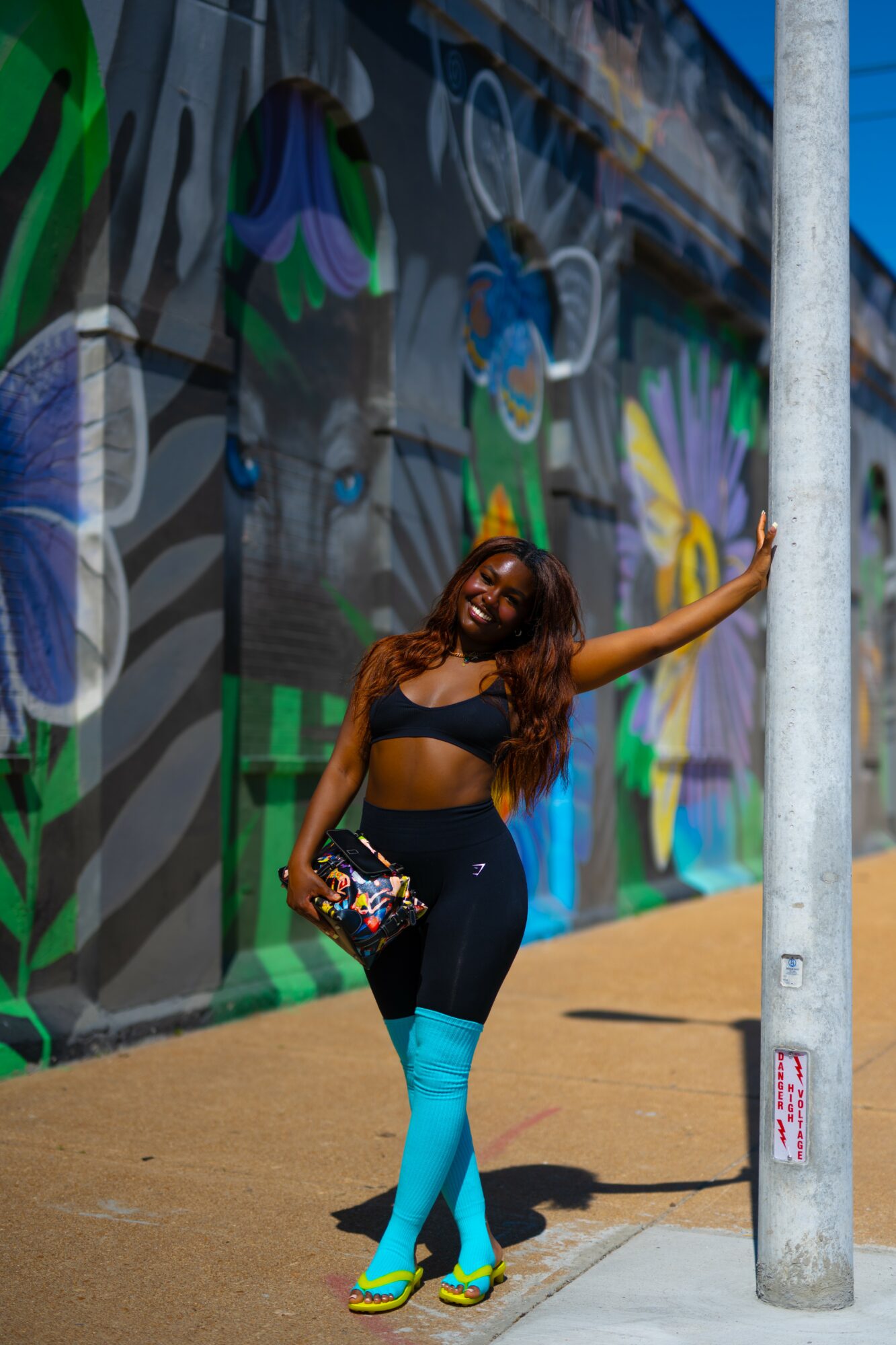 Smiling woman with long hair in a black crop top and blue pants, standing against a colorful mural wall, holding a bag.