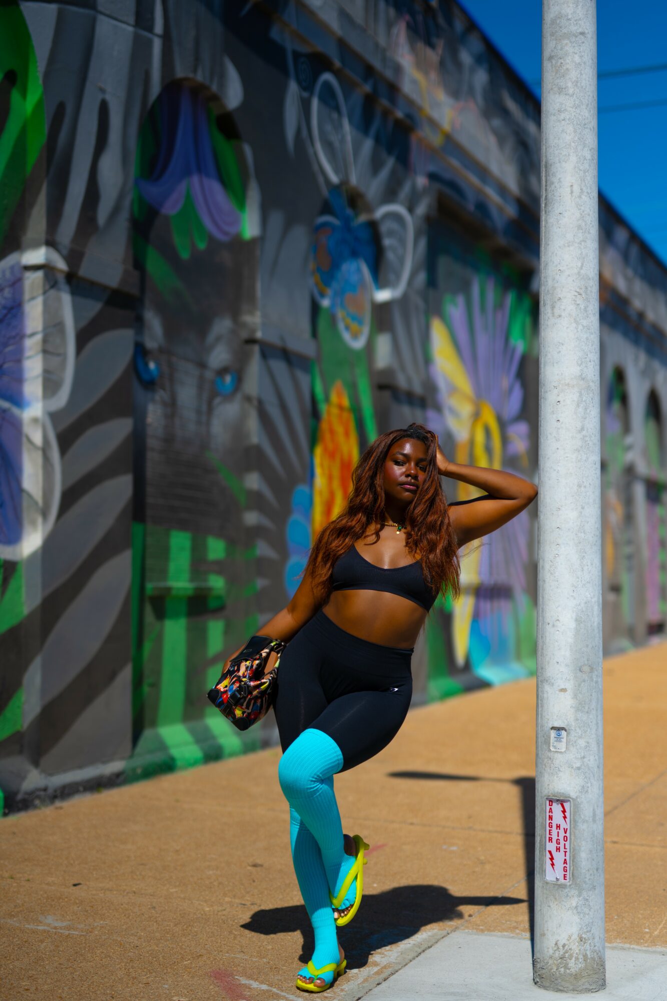 Woman in athletic wear poses outdoors against a colorful graffiti wall, holding a bag, with one hand on her head.