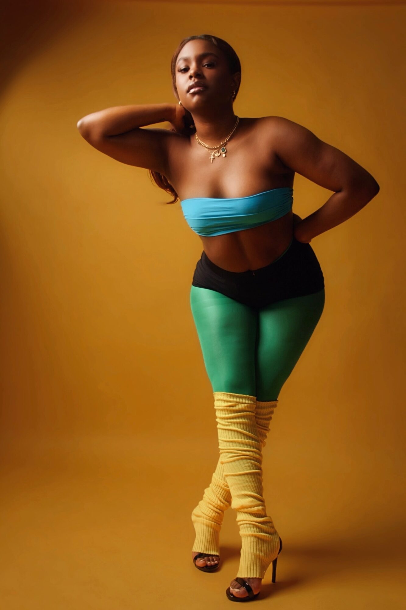 Woman in colorful outfit poses with hand behind neck against yellow background.