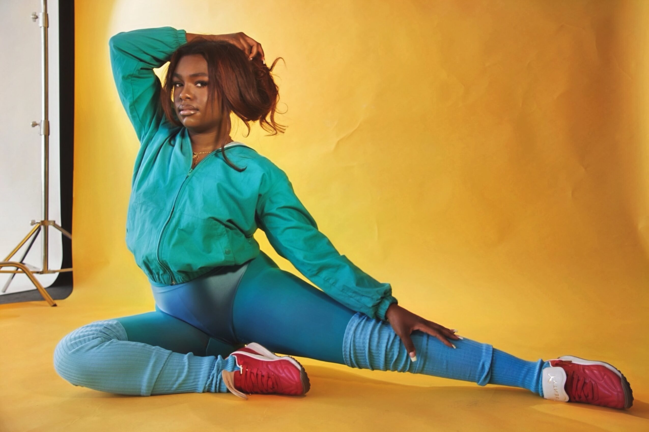 Woman in teal jacket and leggings sitting on yellow background, stretching her leg and resting her hand on her head.