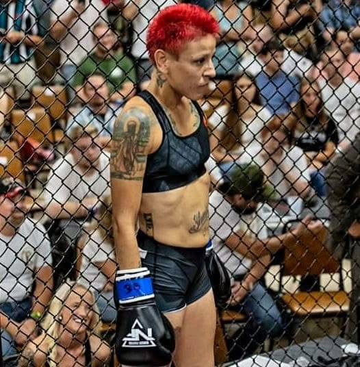 Female mixed martial artist with red hair and tattoos inside a cage, surrounded by spectators.