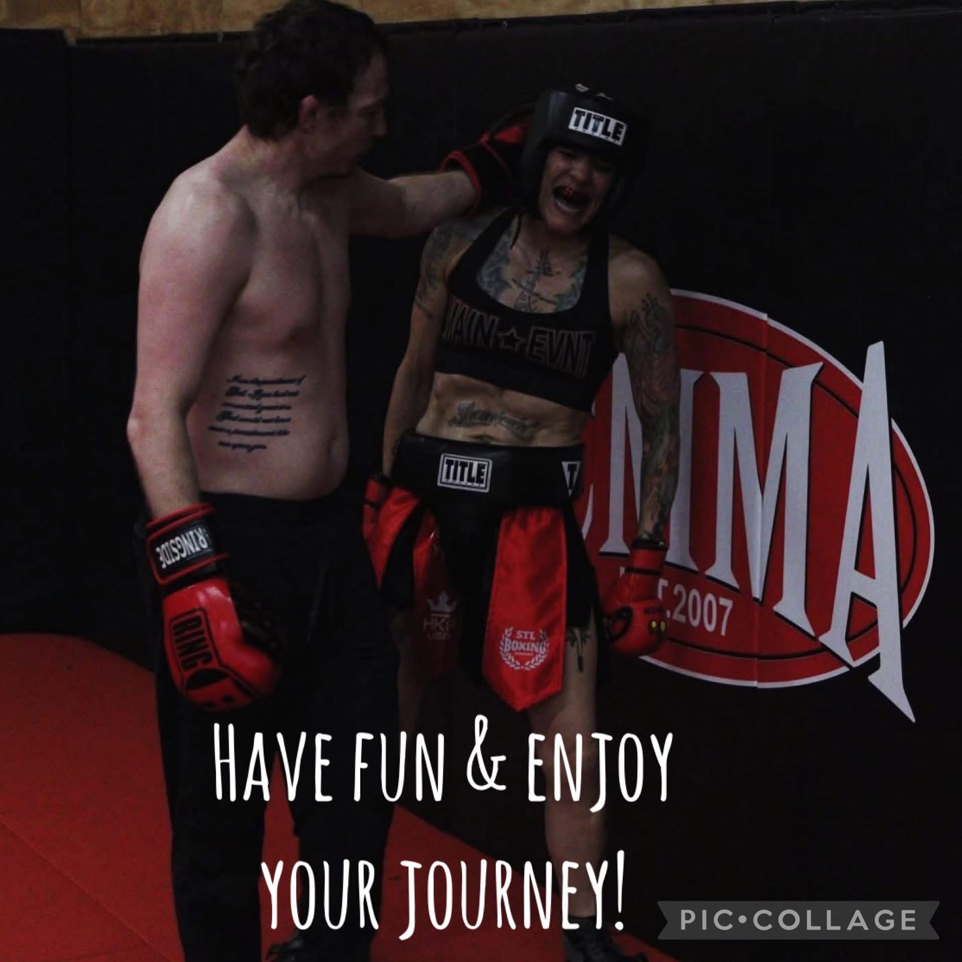 Two people in a gym, one wearing boxing gloves and protective gear, smiling and interacting near a MMA sign.